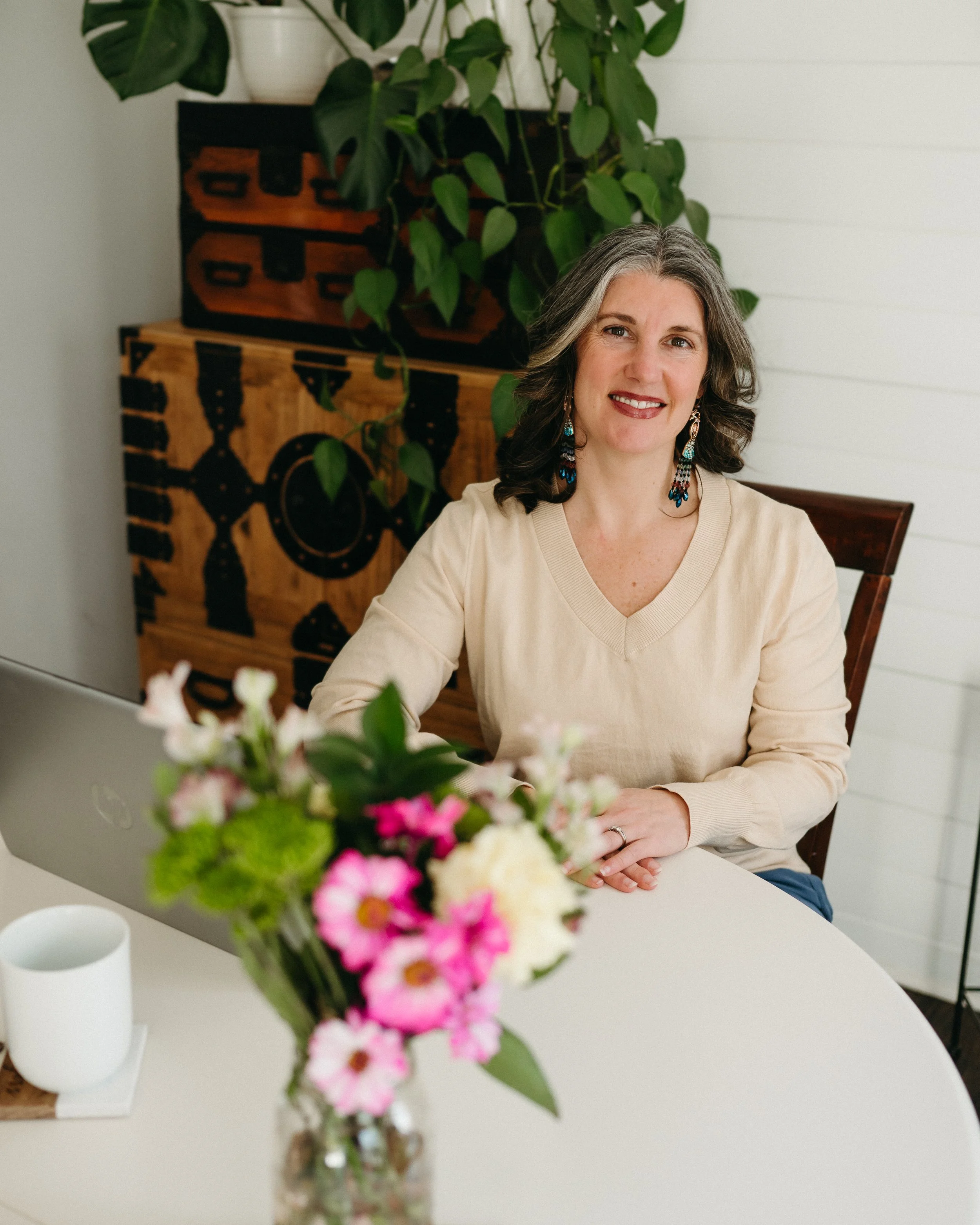 A woman with gray hair and earrings smiling at the camera, sitting at a table with a flower arrangement in the foreground, and a decorated wooden box and green plant in the background.