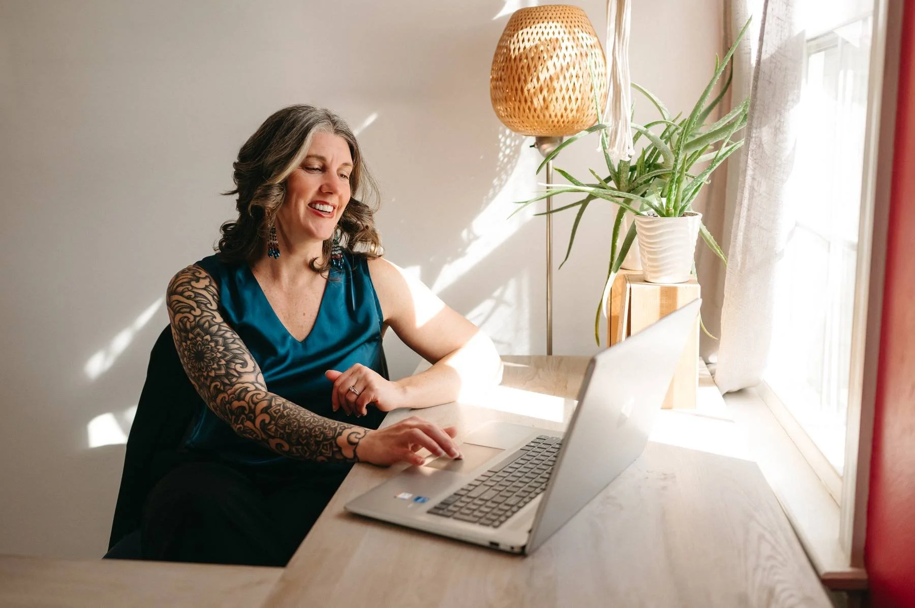 A smiling woman with brown hair and tattoos on her arm, wearing a blue satin top, sitting at a white desk using a laptop. There is a potted plant and a woven lamp on a wooden stand near a window with curtains, sunlight streaming into the room.
