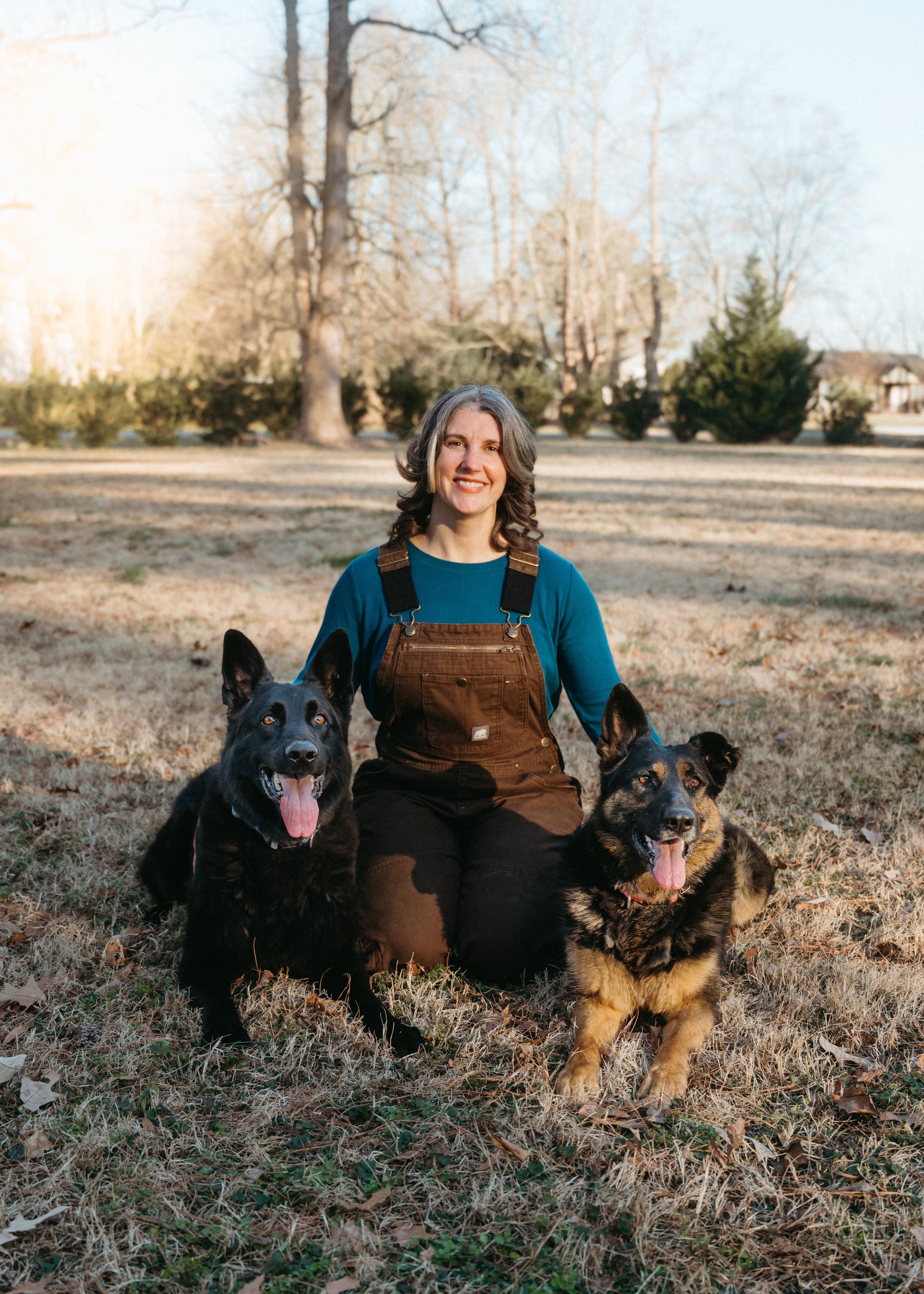 A woman with shoulder-length gray hair, wearing a blue long-sleeve shirt and brown overalls, sitting on grass in a park with two dogs, one black and one black and tan, both with their tongues out, in front of large trees with bare branches during late fall or winter.