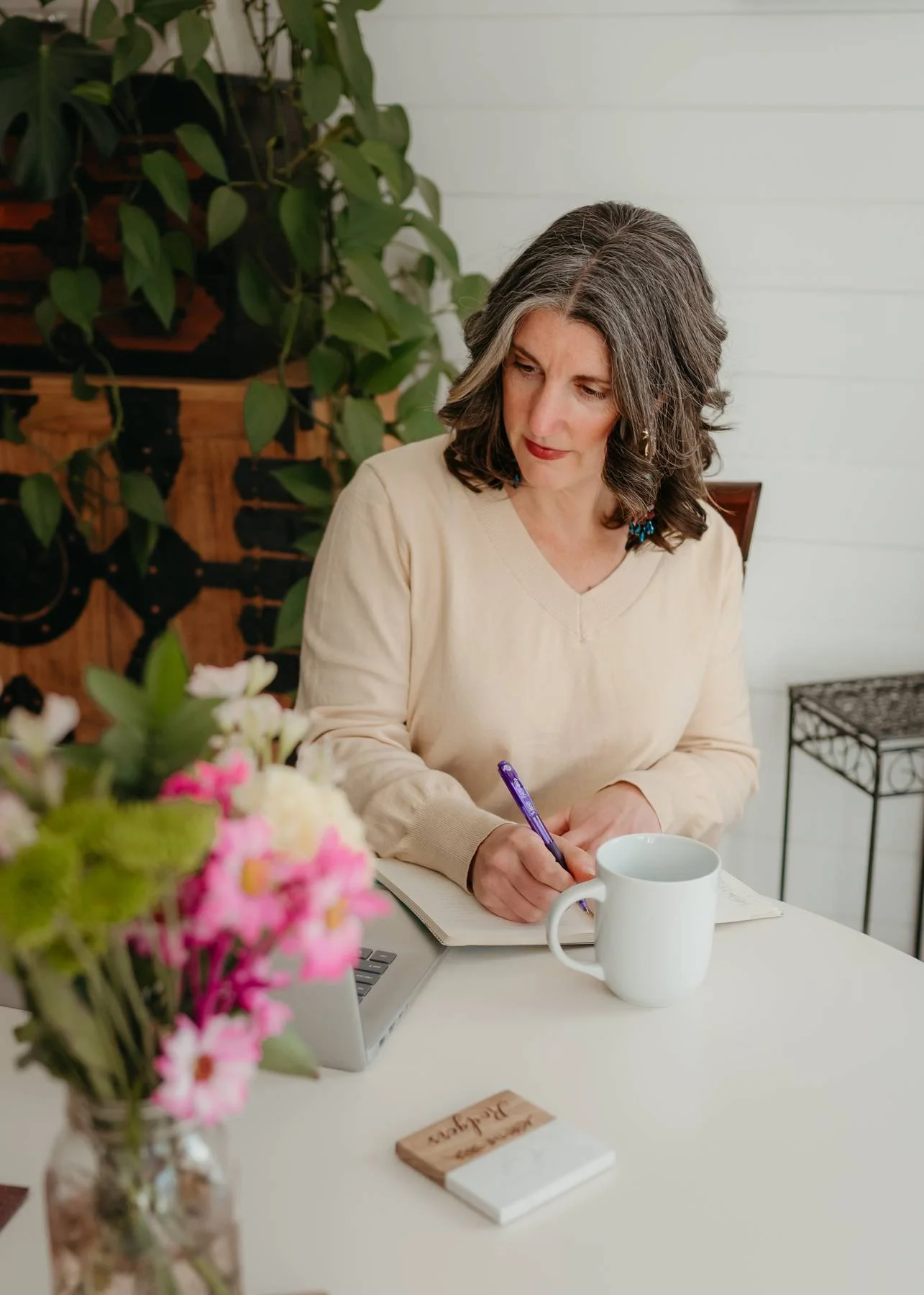 A woman with gray and dark wavy hair wearing a cream sweater, sitting at a white table writing in a notebook with a purple pen, with a white mug in front of her, a bouquet of pink and white flowers, a laptop, and some notes on the table. There are green plants and a black metal stand in the background.