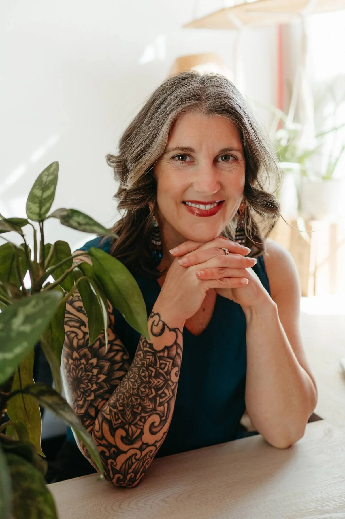 Smiling woman with gray hair and tattoos on her arms, sitting at a table with her hands clasped under her chin, near potted plant.
