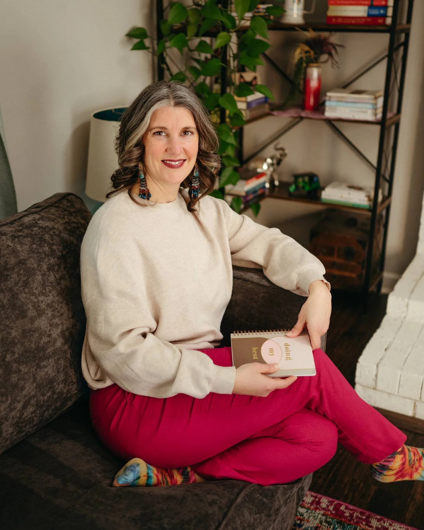 A woman with wavy gray hair, wearing a beige sweater and red pants, sitting on a brown couch in a cozy room. She is smiling while holding a notebook that says "doing my best." Behind her, there is a bookshelf with books, plants, and decorative items, and a lamp with a white shade.