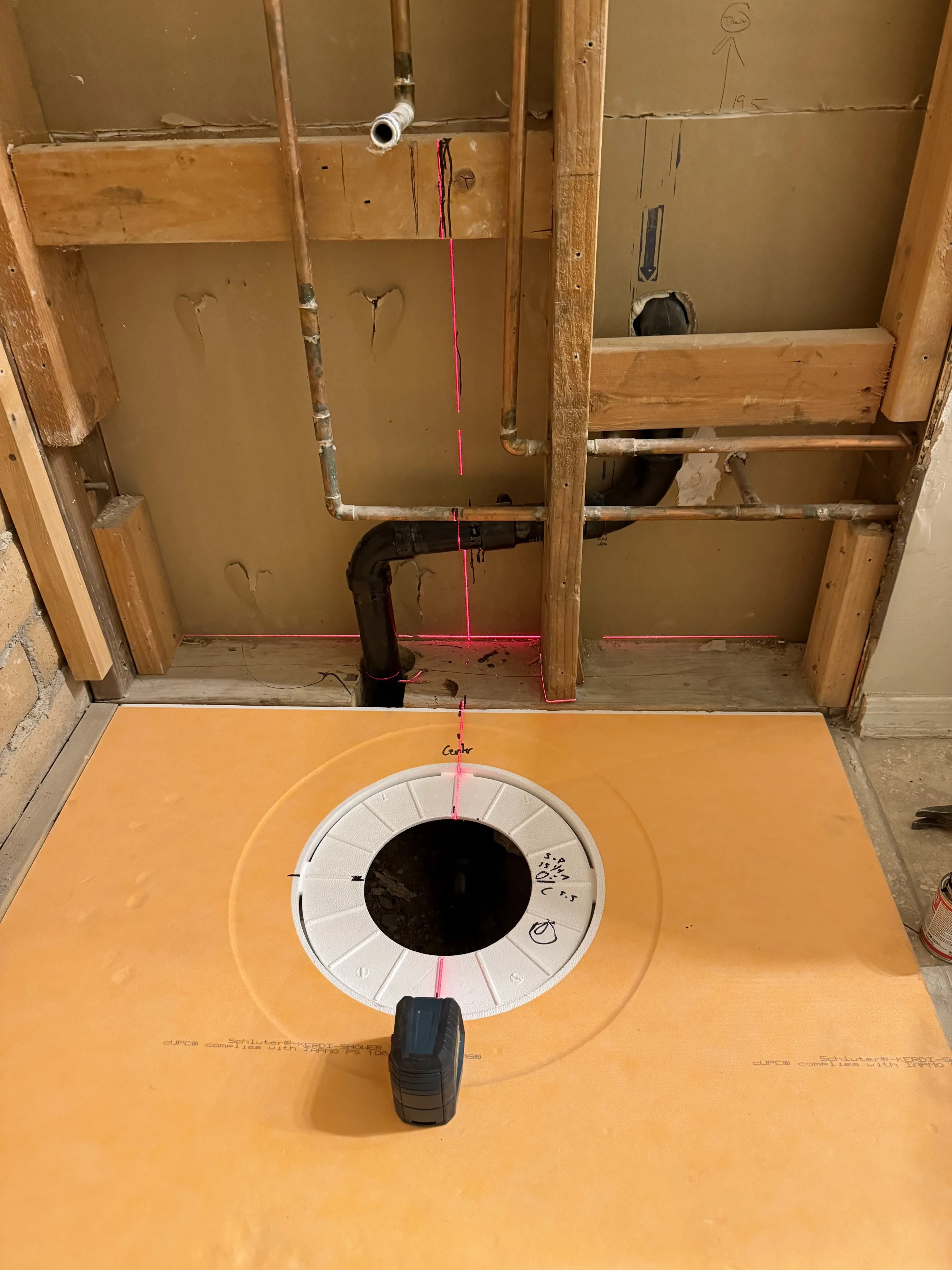 Construction site showing plumbing installation with laser level and drywall framing, with a circular cutout in orange flooring for pipework.