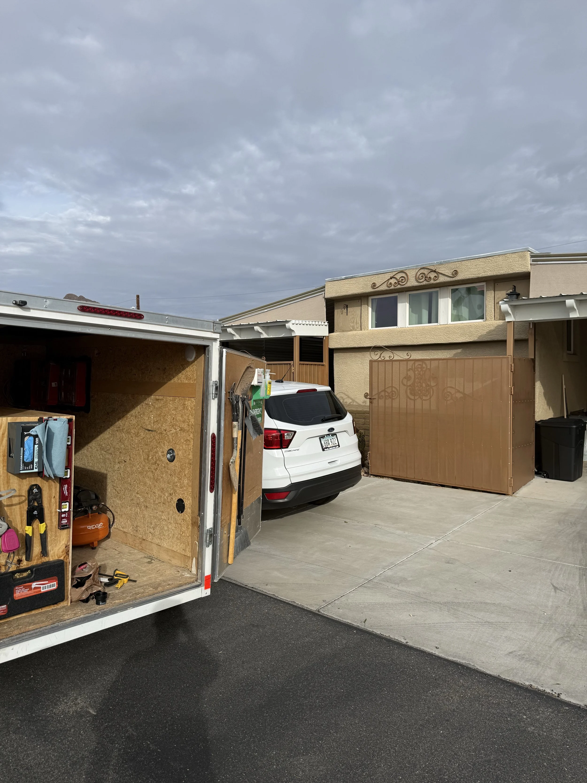 Residential driveway with a white SUV and a trailer, a brown metal gate, a trash bin, and a two-story house with beige stucco walls and decorative ironwork, under a cloudy sky.