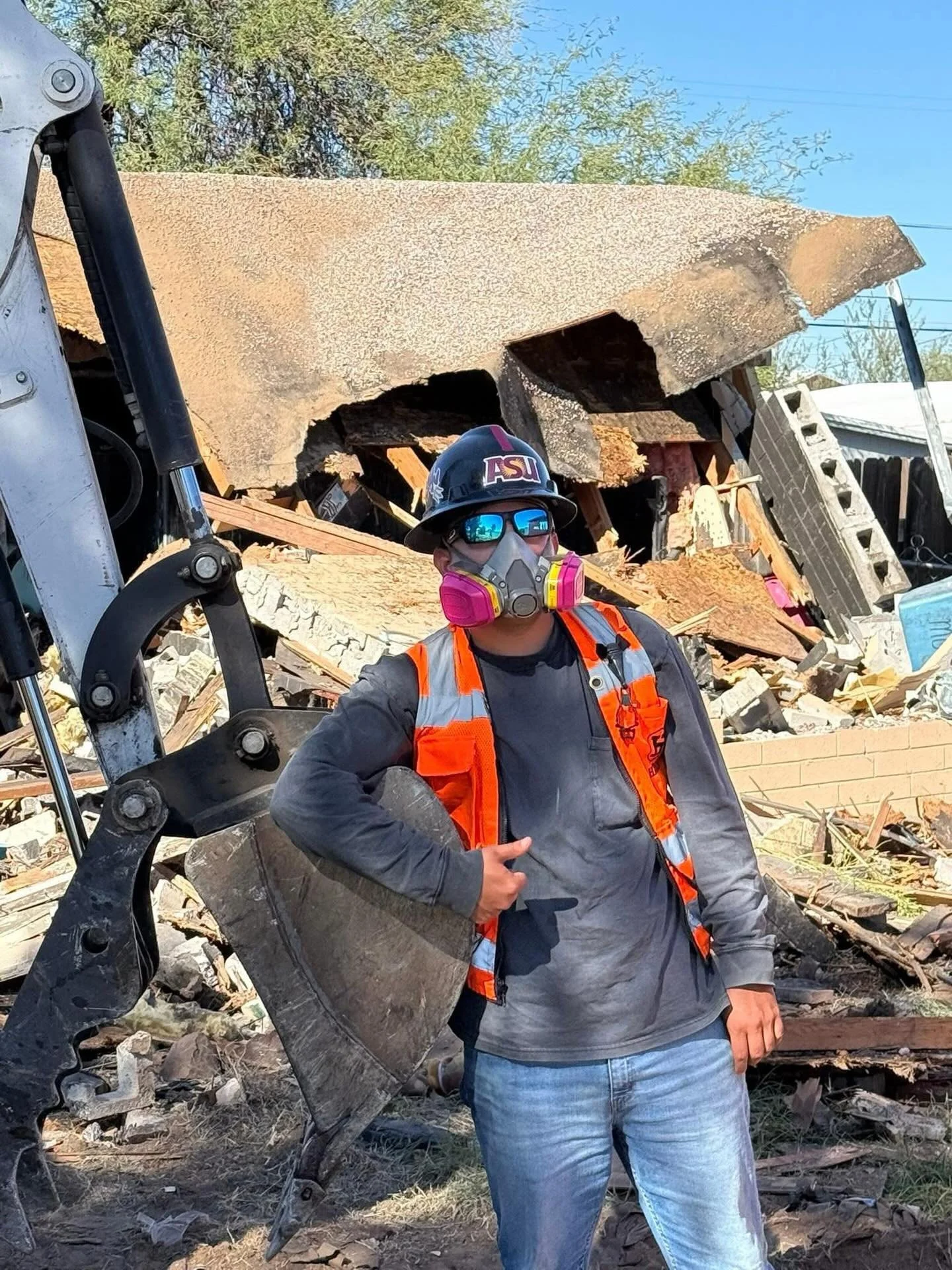 A person wearing a black hard hat, sunglasses, a respirator mask, an orange safety vest, a gray long-sleeve shirt, and jeans, standing in front of a collapsed building with debris and plywood. The person is giving a thumbs-up and holding a piece of debris.