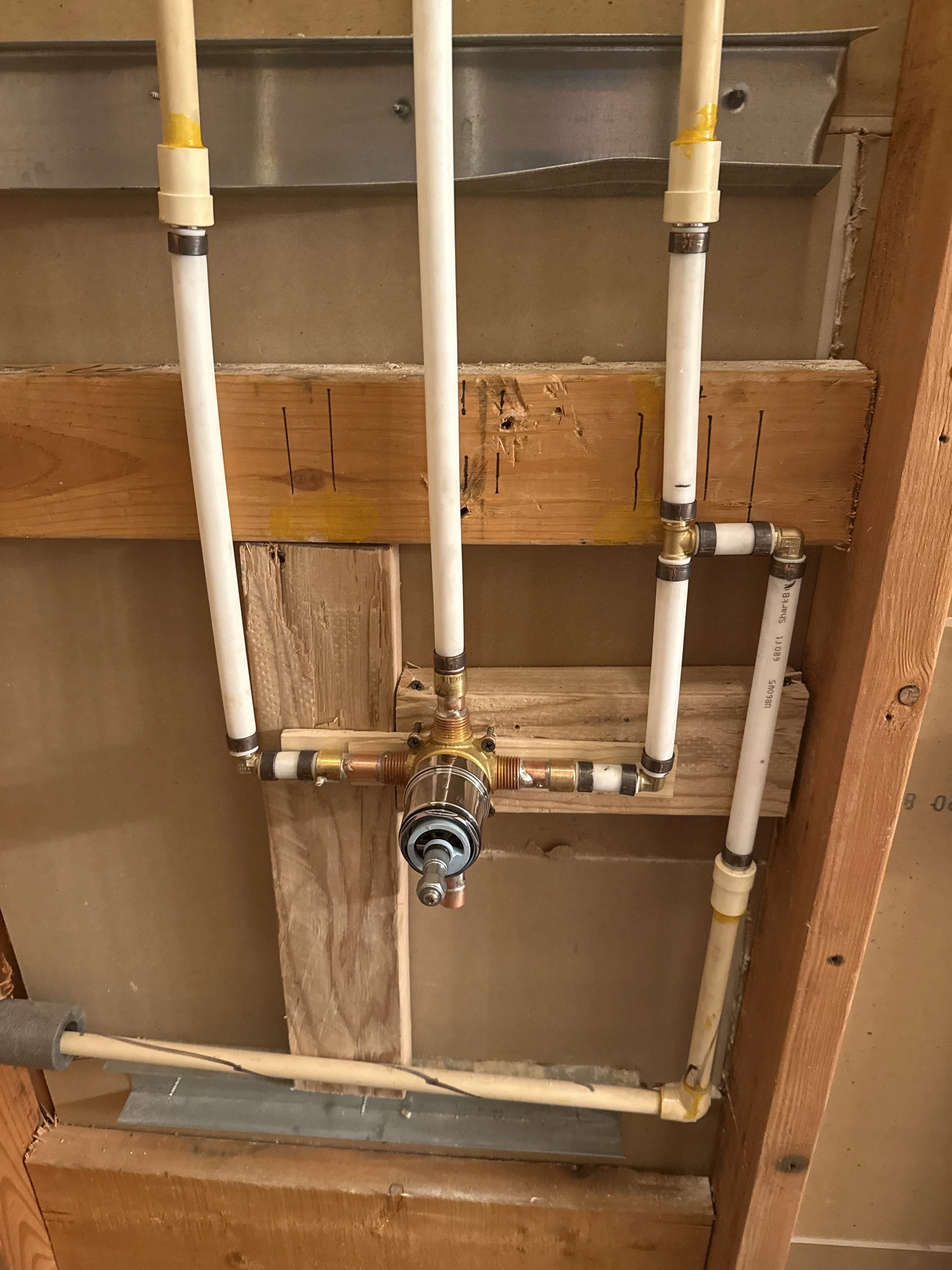 A plumbing installation with white PVC pipes connected to a brass valve assembly, mounted on unfinished wooden studs inside a wall cavity.