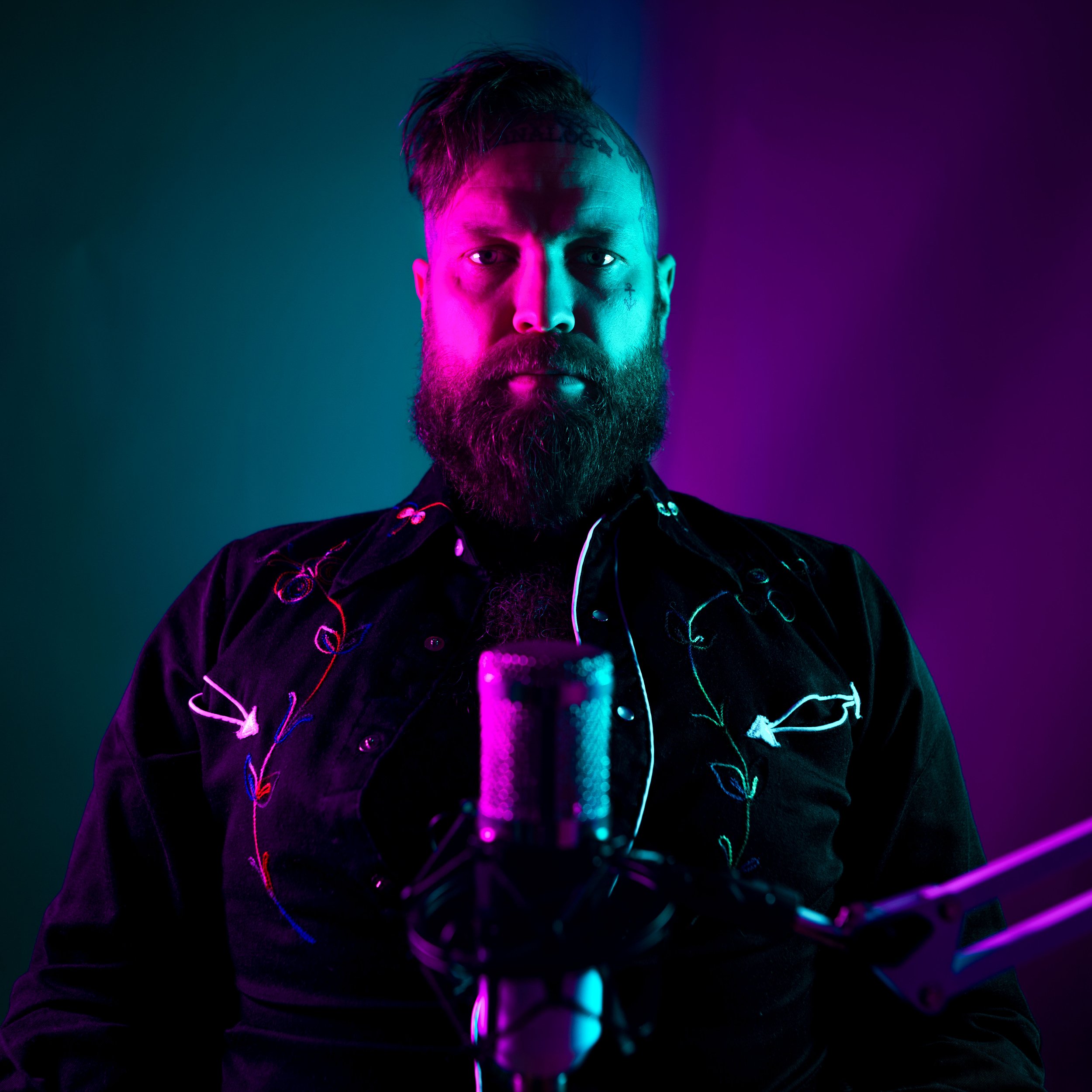 Bearded Reggie Grey with a mohawk hairstyle sits in front of a microphone, illuminated by vibrant purple and teal lighting. Hipster as heck. Way too cool.