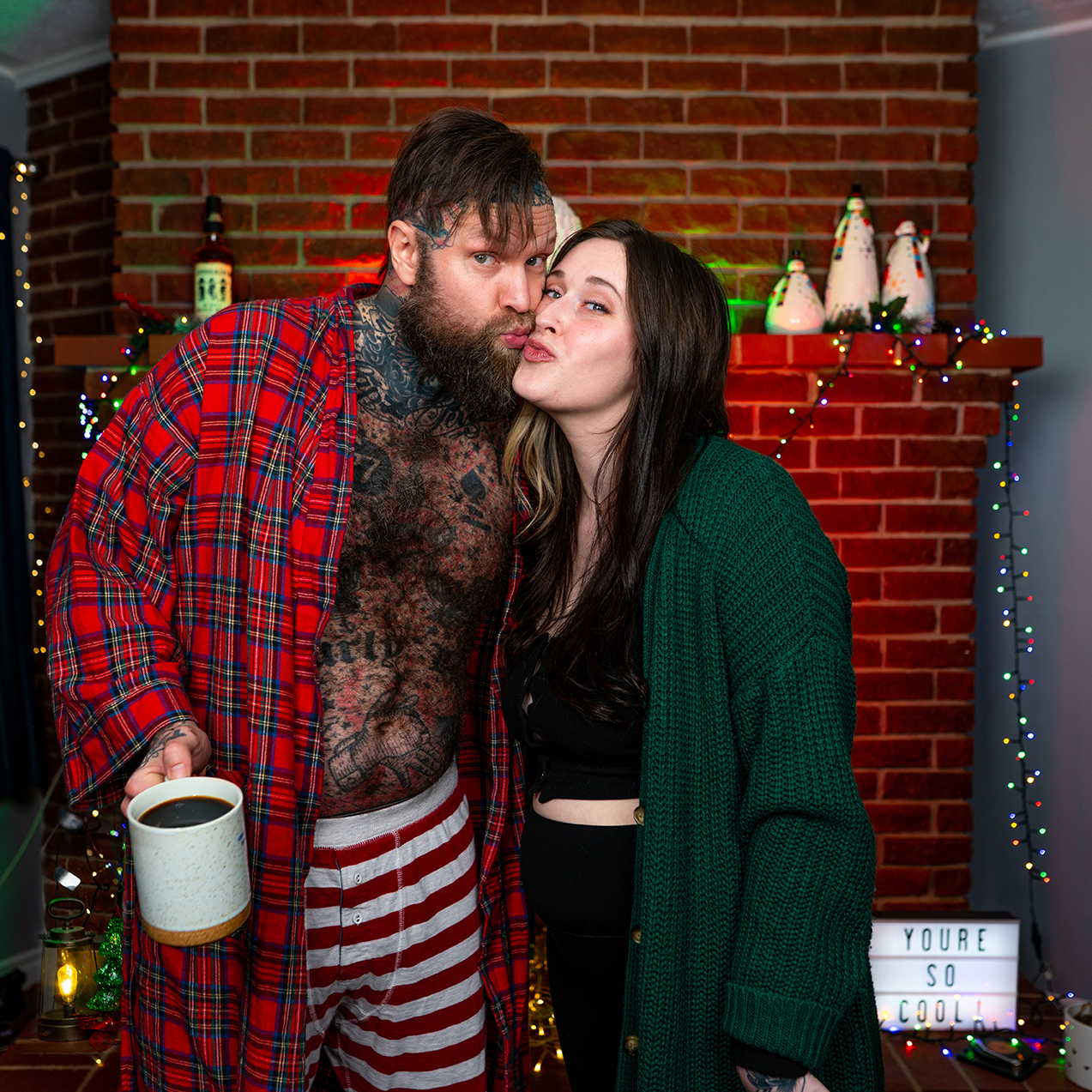 Reggie Grey and his wife in their pyjamas, pose with a coffee infront of their fireplace. There is a sign that says "You're So Cool!" in the background. They love each other, proven by a side kiss.