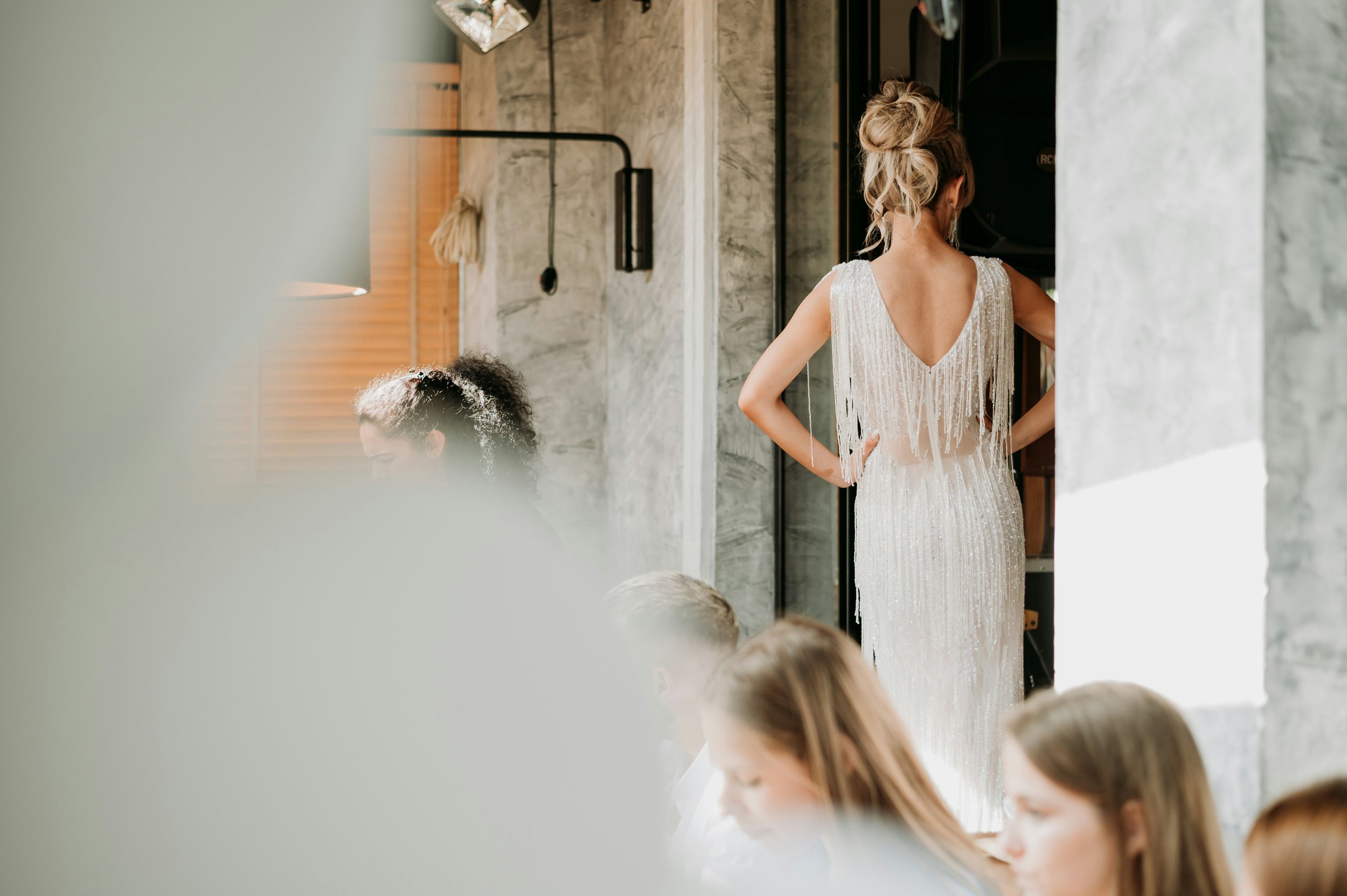 A woman in a white, beaded dress with fringe detail stands with her back to the camera, surrounded by other women seated indoors.