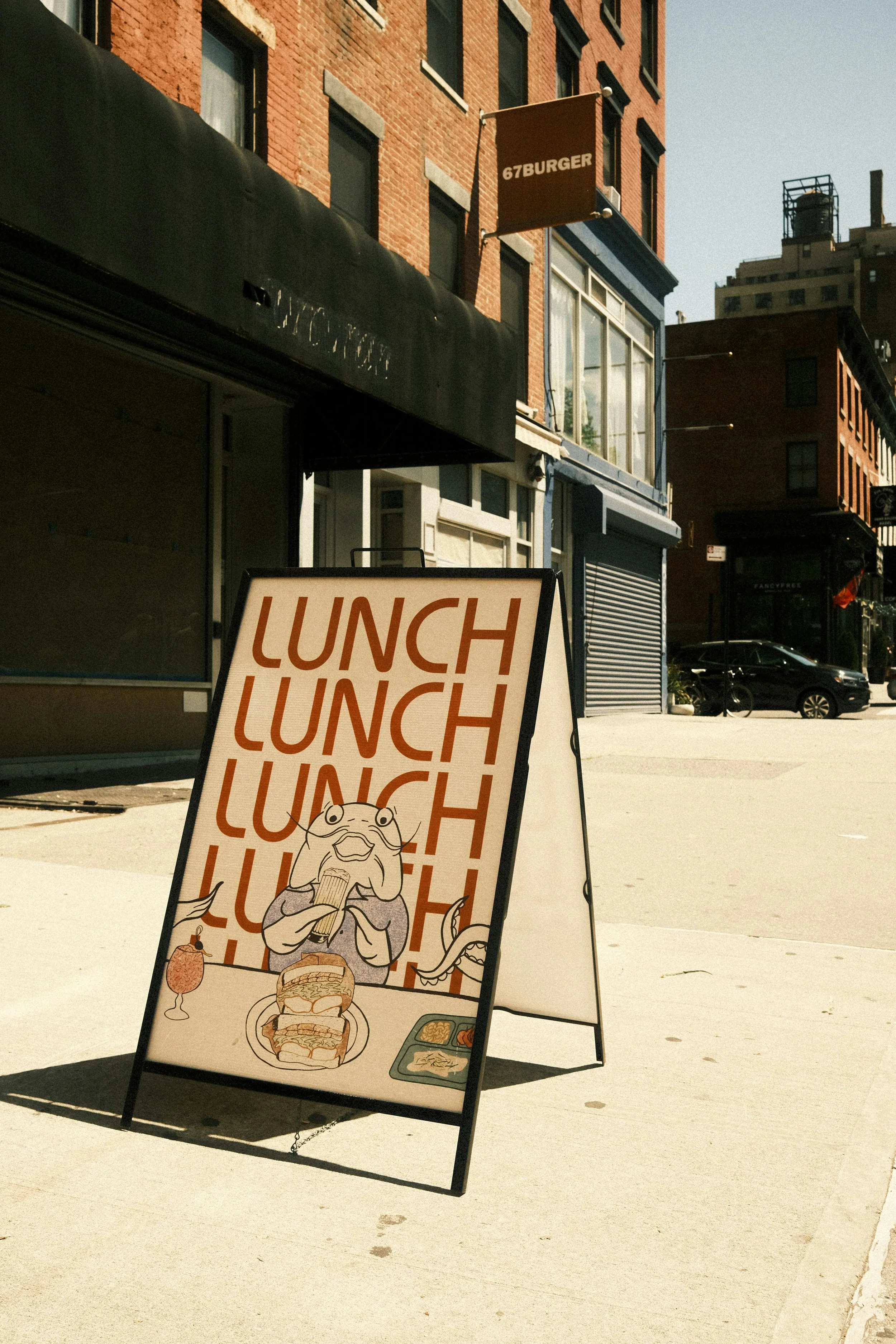 A sidewalk sign advertising lunch outside a restaurant with the text 'LUNCH' repeated multiple times, an illustration of a frog eating, a burger, two drinks, and food trays.
