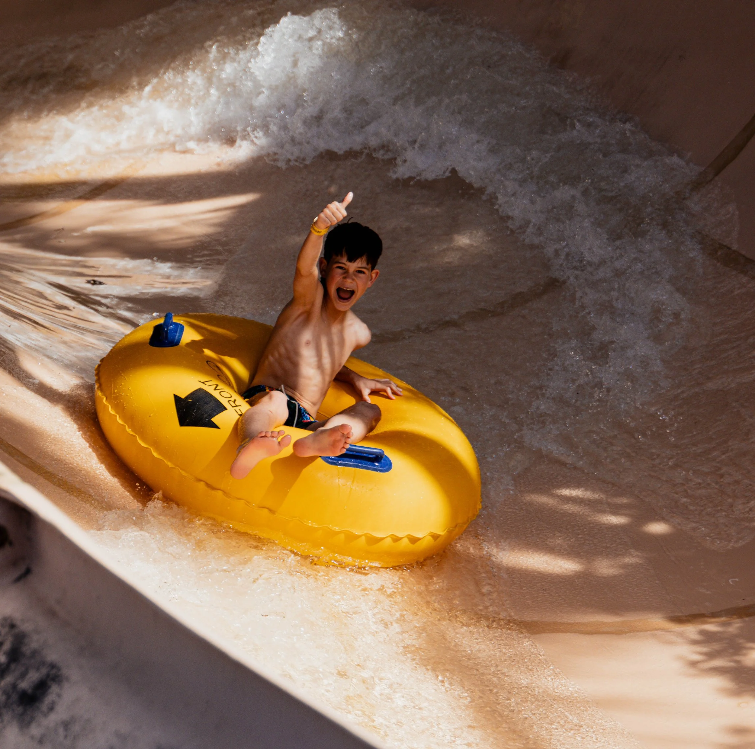 A young boy in swim trunksjoyfully riding a yellow inflatable raft down a water slide.
