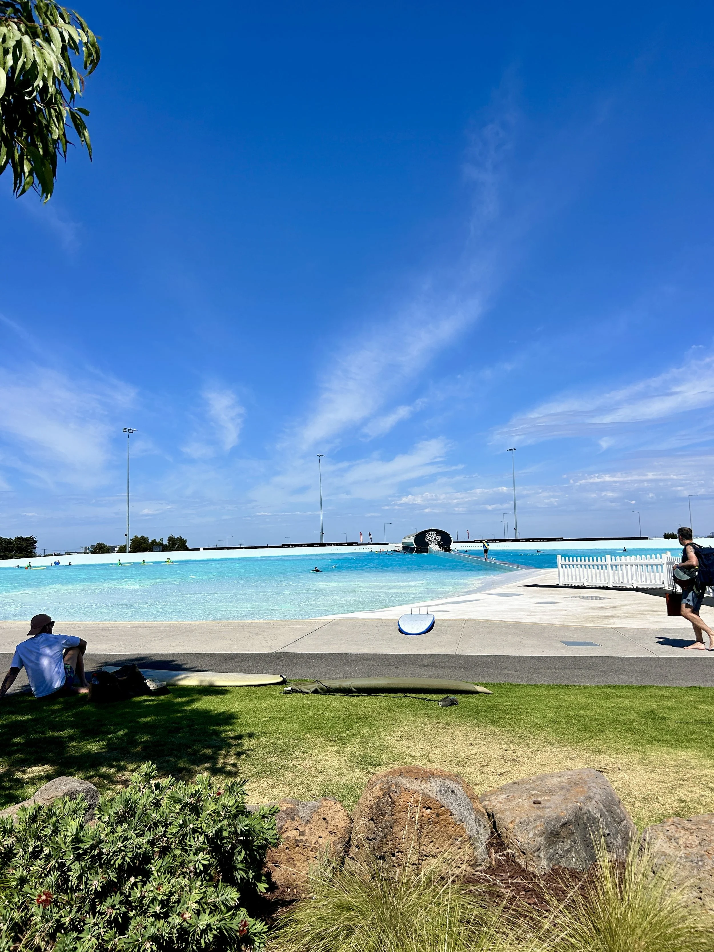 People sitting and walking near an outdoor swimming pool with a water slide, under a clear blue sky, surrounded by grass, rocks, and some greenery.