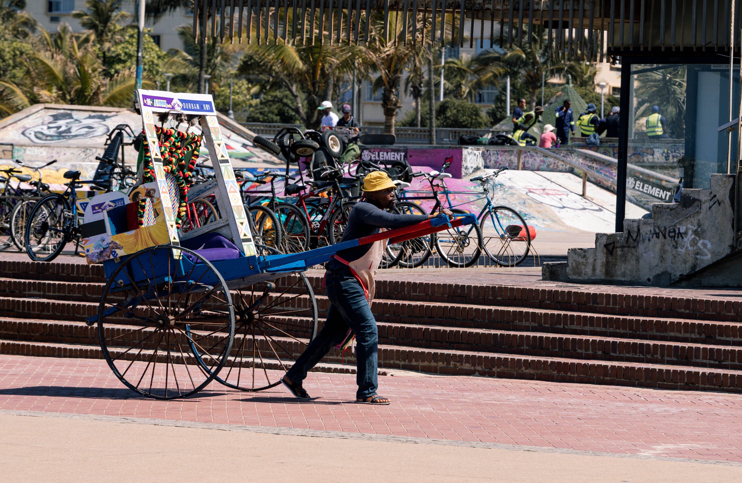 A man pushing a colorful cart with a small platform on top, walking along a brick-paved area in a skatepark, with bicycles and graffiti-covered ramps in the background.