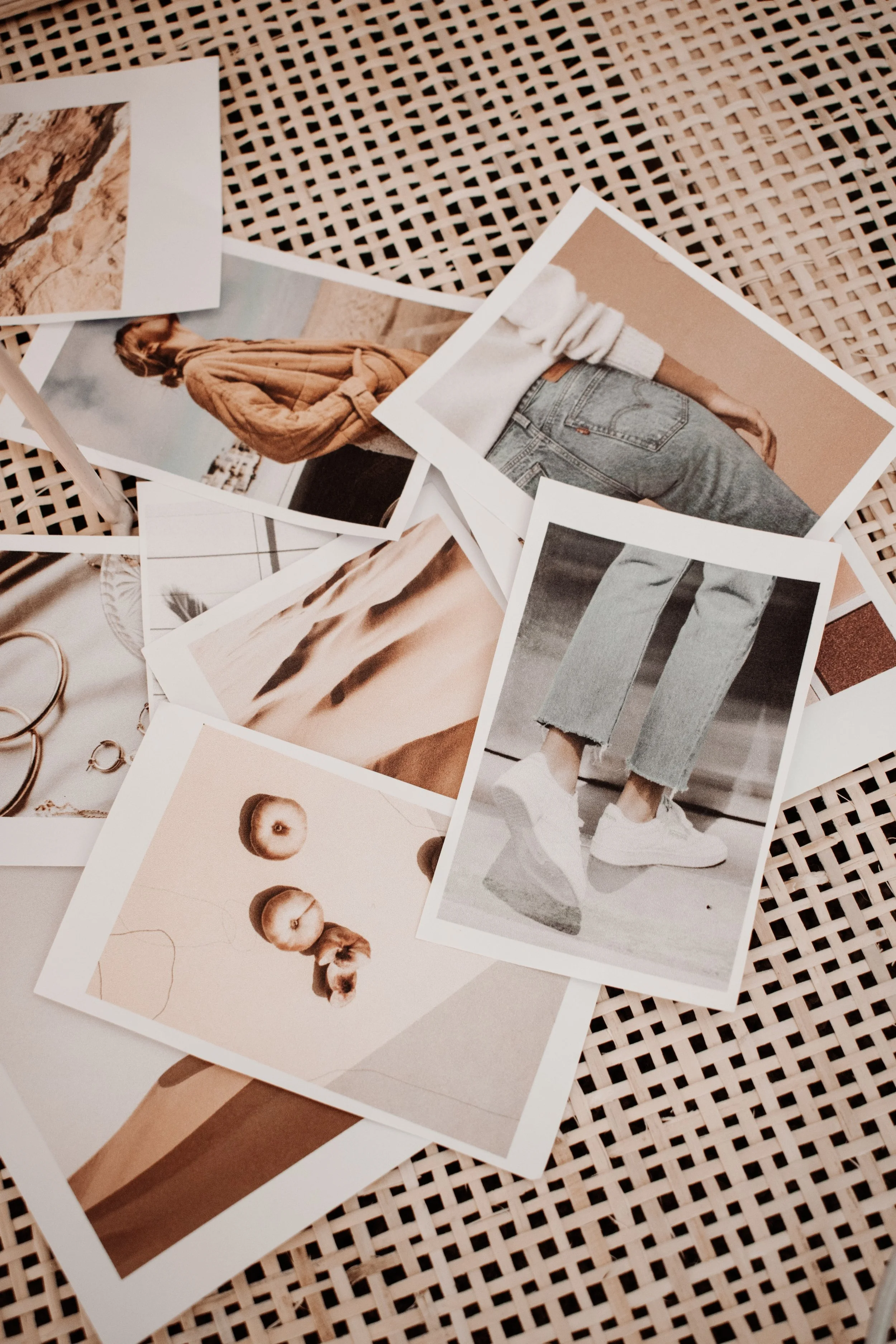 A collection of printed photographs scattered on a woven surface, including images of a woman in a tan coat, jeans and sneakers, desert landscapes, jewelry, and close-ups of fruit.