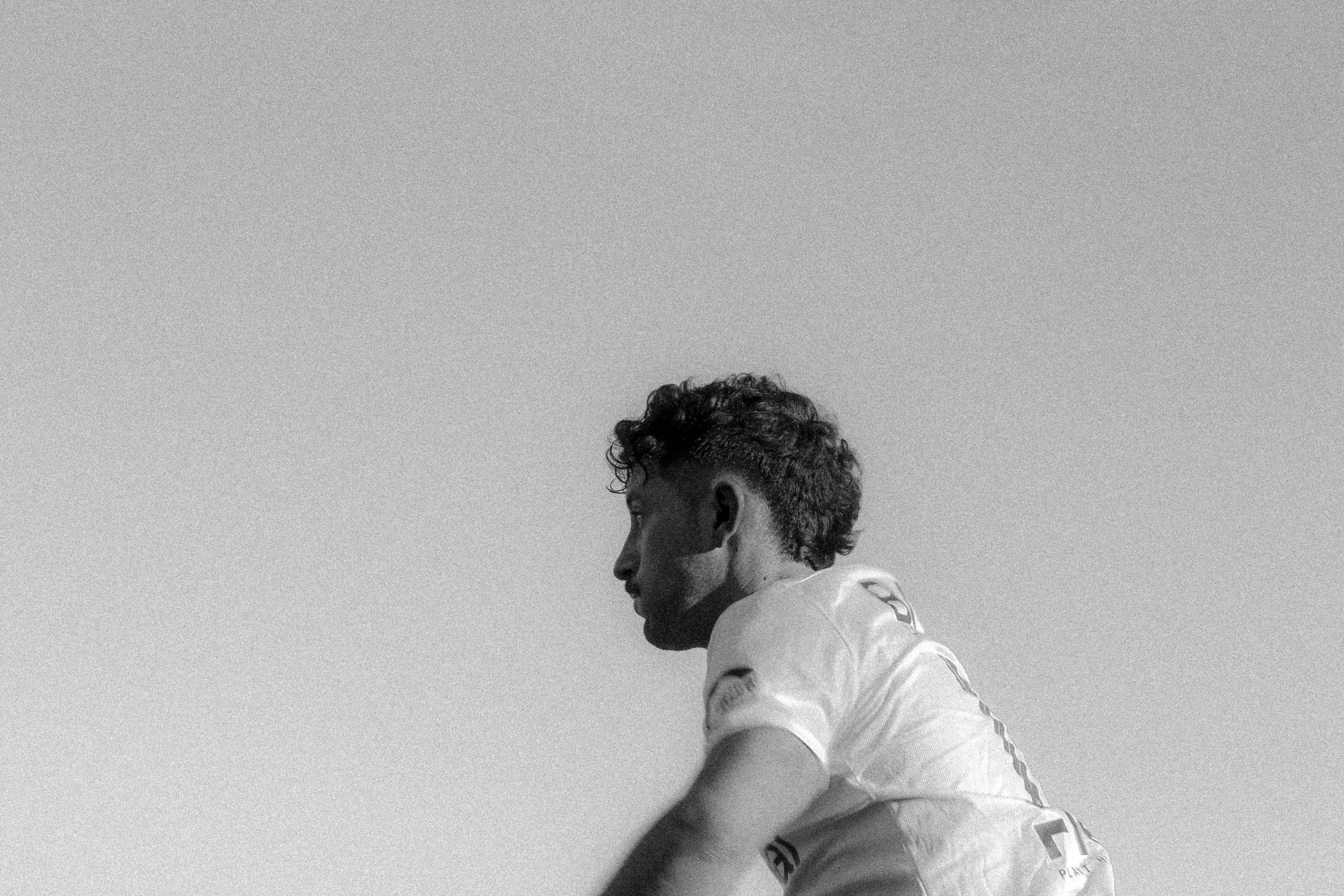 Side profile of a young male athlete with curly hair, wearing a sports jersey, outdoors against a clear sky.