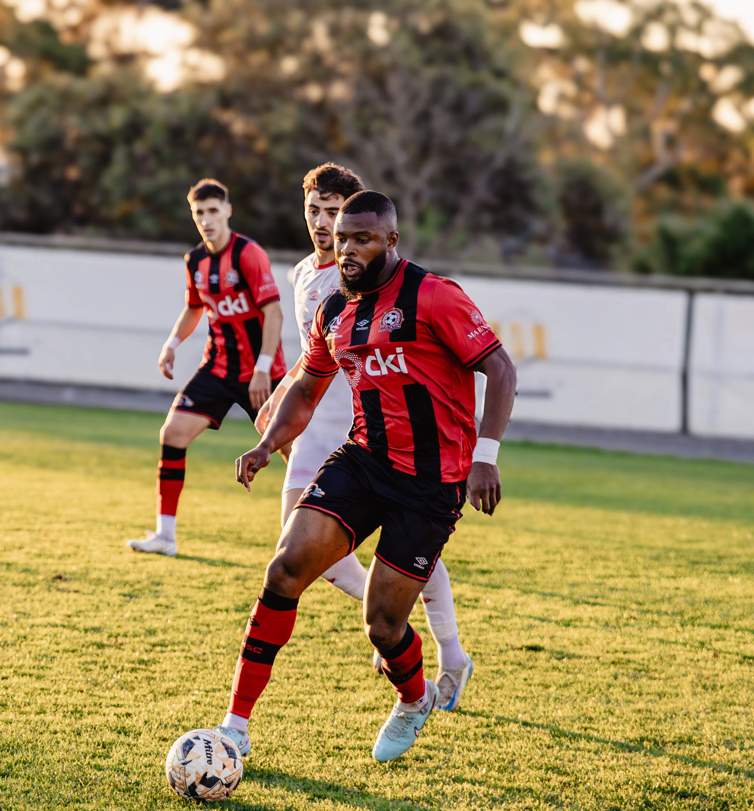 Three soccer players on a field during a game, with two wearing red and black striped uniforms and one in white, all focused on controlling the soccer ball.
