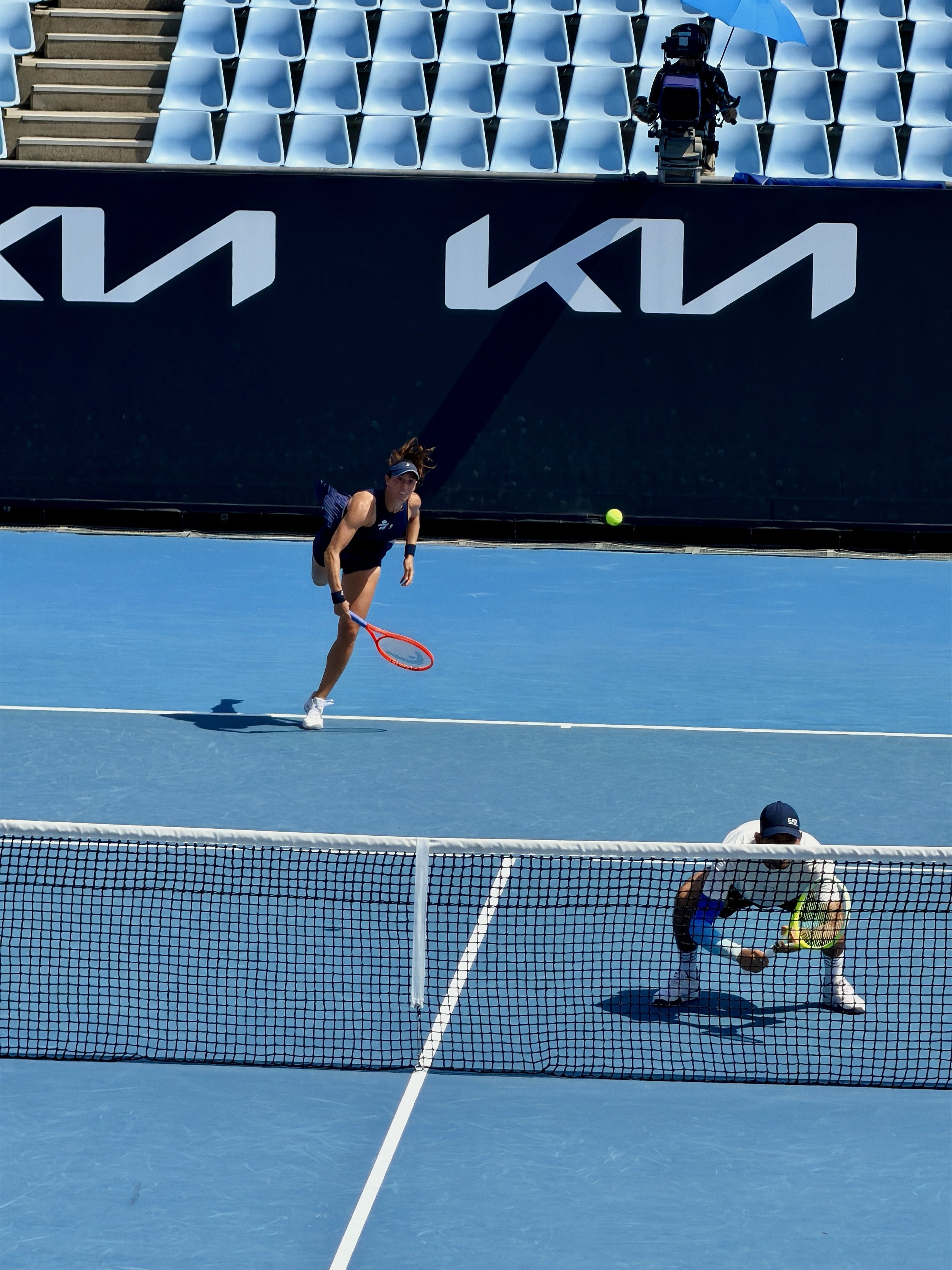 A woman playing tennis on a blue court with a tennis ball in the air, a man crouched behind the net, and empty blue seats in the background.