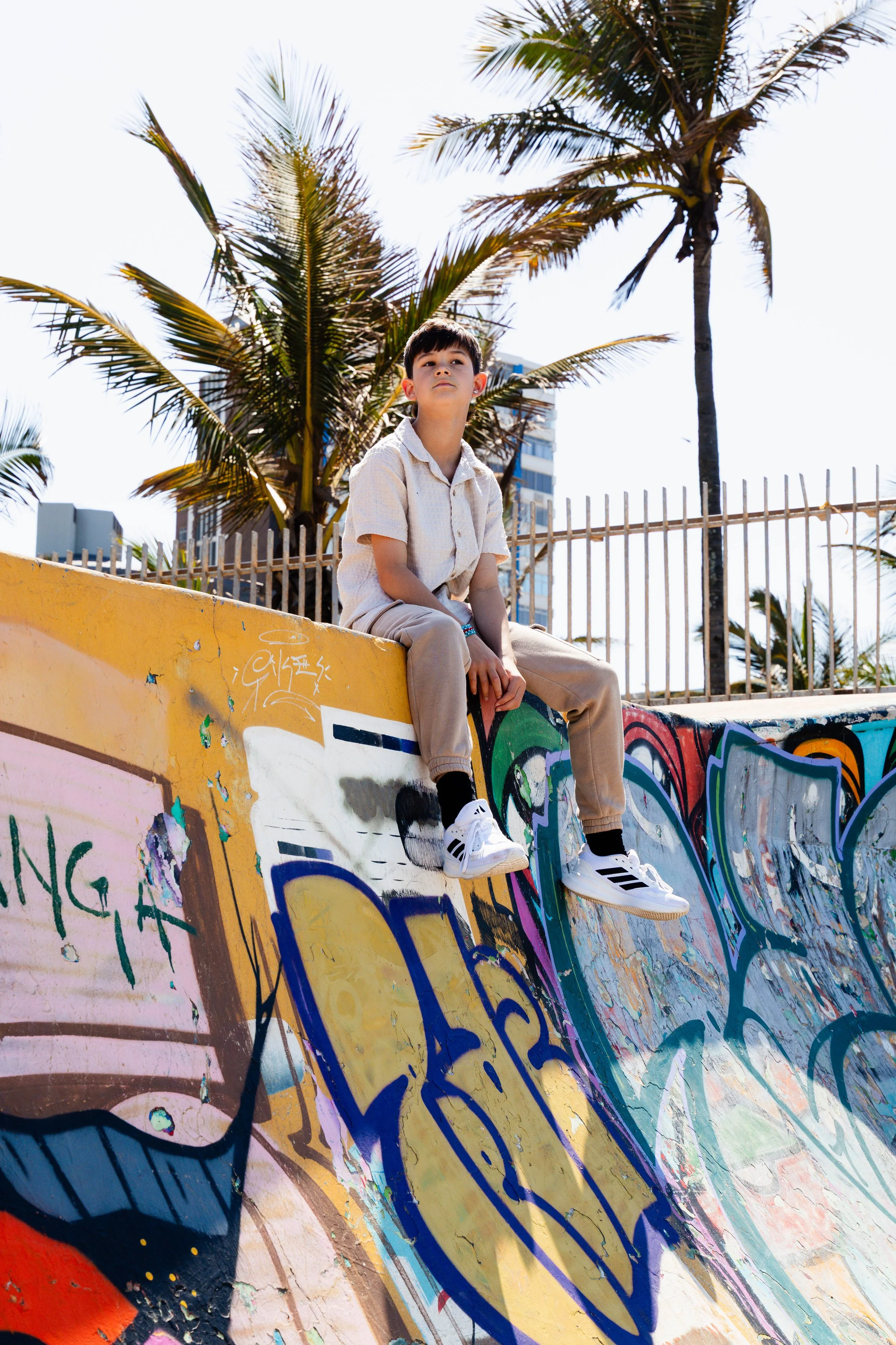 Young boy sitting on a colorful graffiti-covered skatepark ramp with palm trees and a fence in the background under a clear sky.