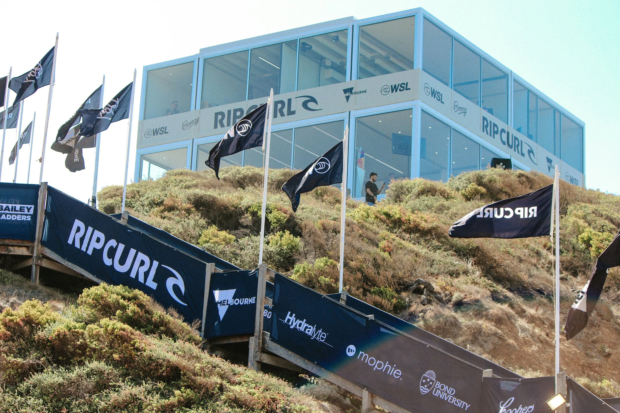 A hillside with flagged banners and a glass-walled building on top, displaying the words 'Rip Curl' and 'WSL'.
