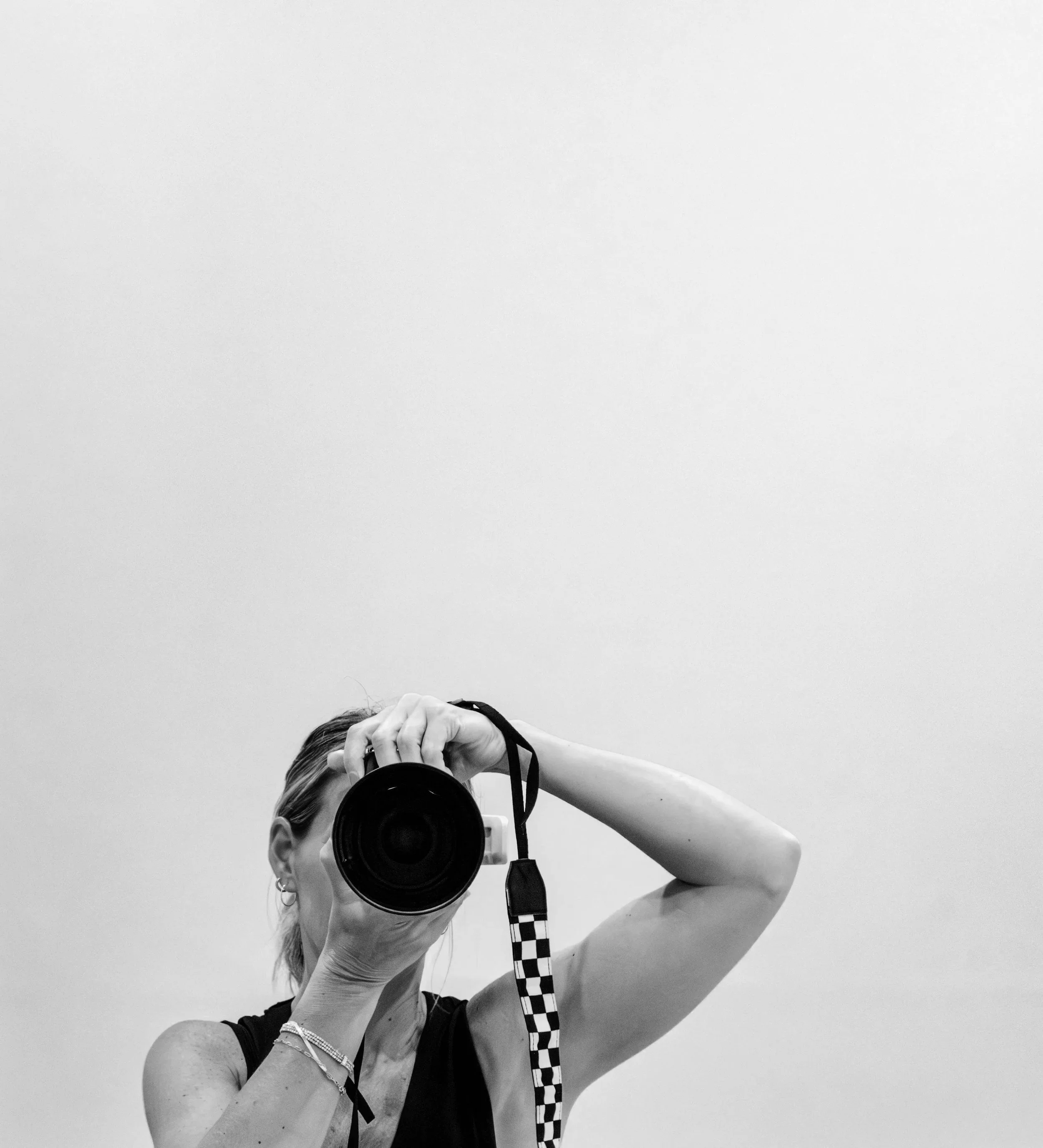 A woman taking a photo of herself in a mirror with a digital camera, wearing a sleeveless top and jewelry, with a plain white background.