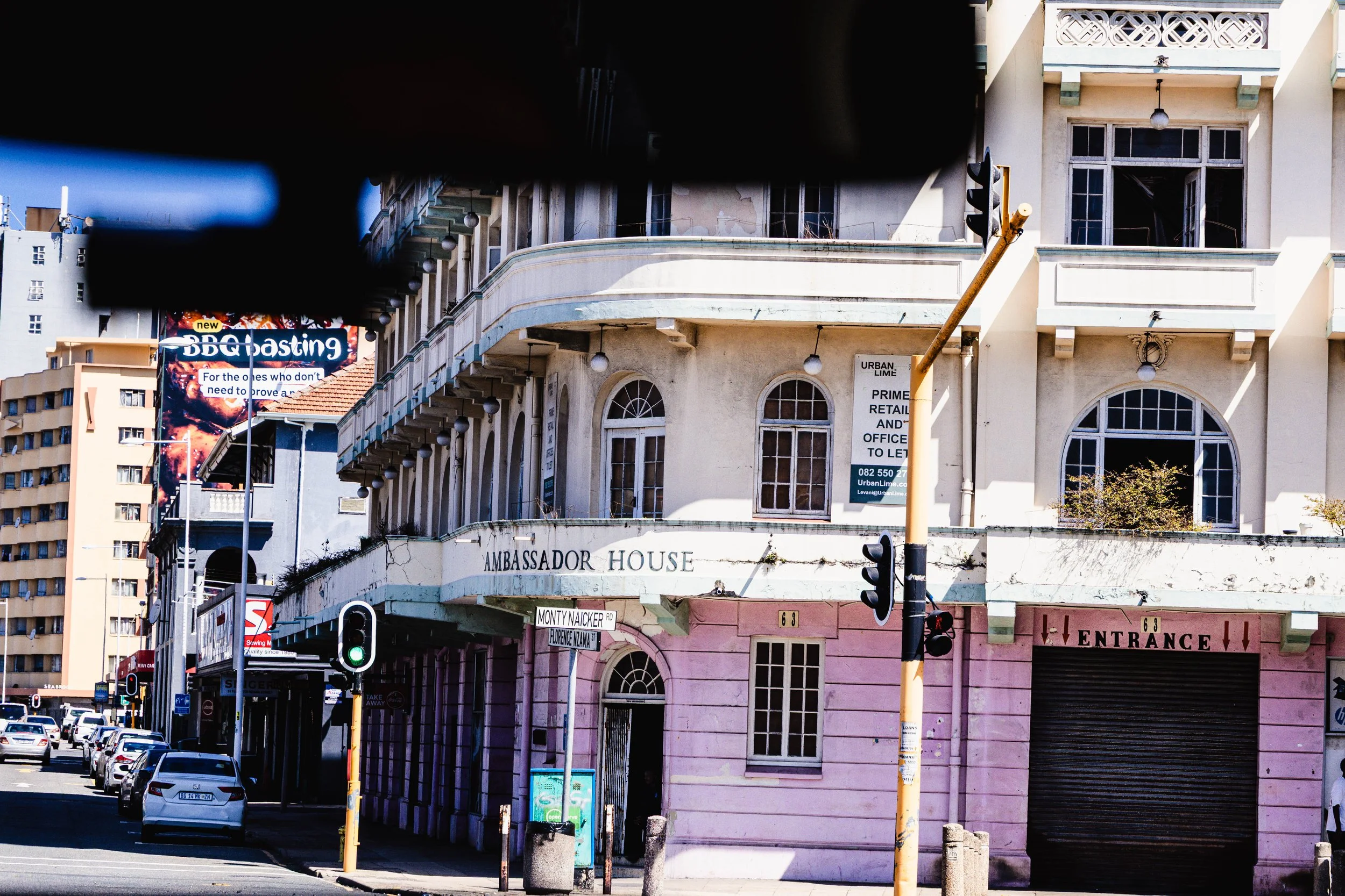 A street scene featuring the Ambassador House building with a pink lower section and white upper section, with signs indicating it is for lease. There are multiple parked cars and traffic lights, and a billboard advertising a BBQasting event in the b