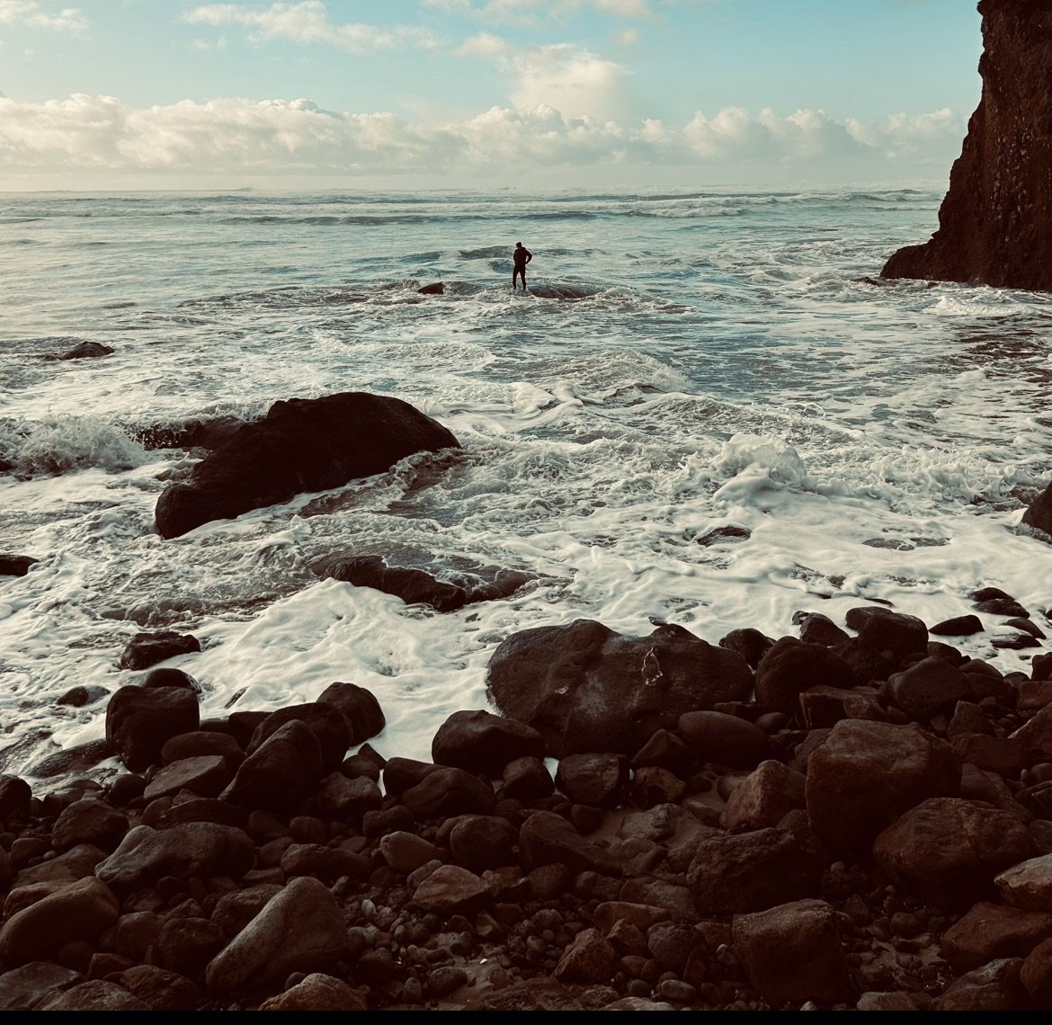Personal inspirational reference of a person standing on rocks at the edge of the ocean, with waves crashing against the rocks and a cloudy sky overhead.