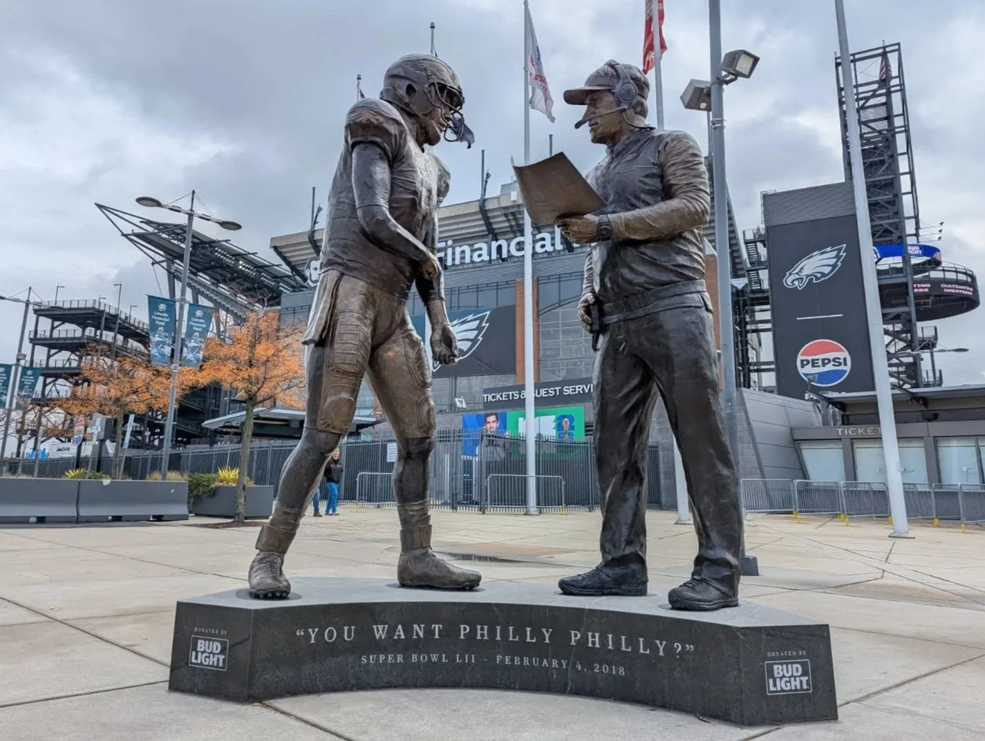Bronze statue of a football player and a coach outside Lincoln Financial Field in Philadelphia, Pennsylvania. The statue depicts a football player in uniform and a coach in a cap, with the coach reading from a clipboard. The background shows the stadium entrance with banners and signs for the Philadelphia Eagles.