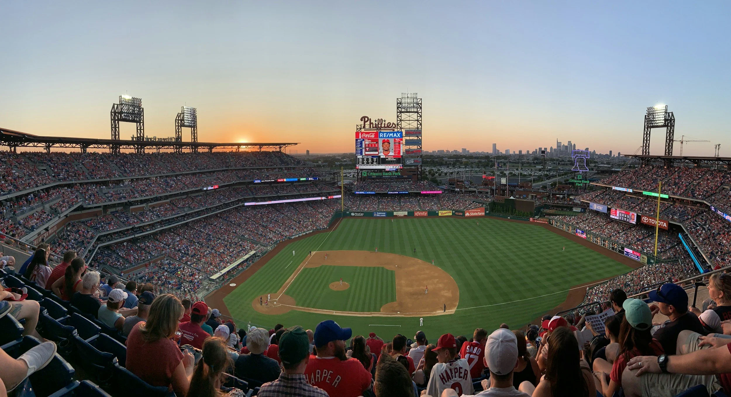 Photograph of a baseball game at Citizens Bank Park stadium during sunset, with fans in the stands, players on the field, and the city skyline in the background.