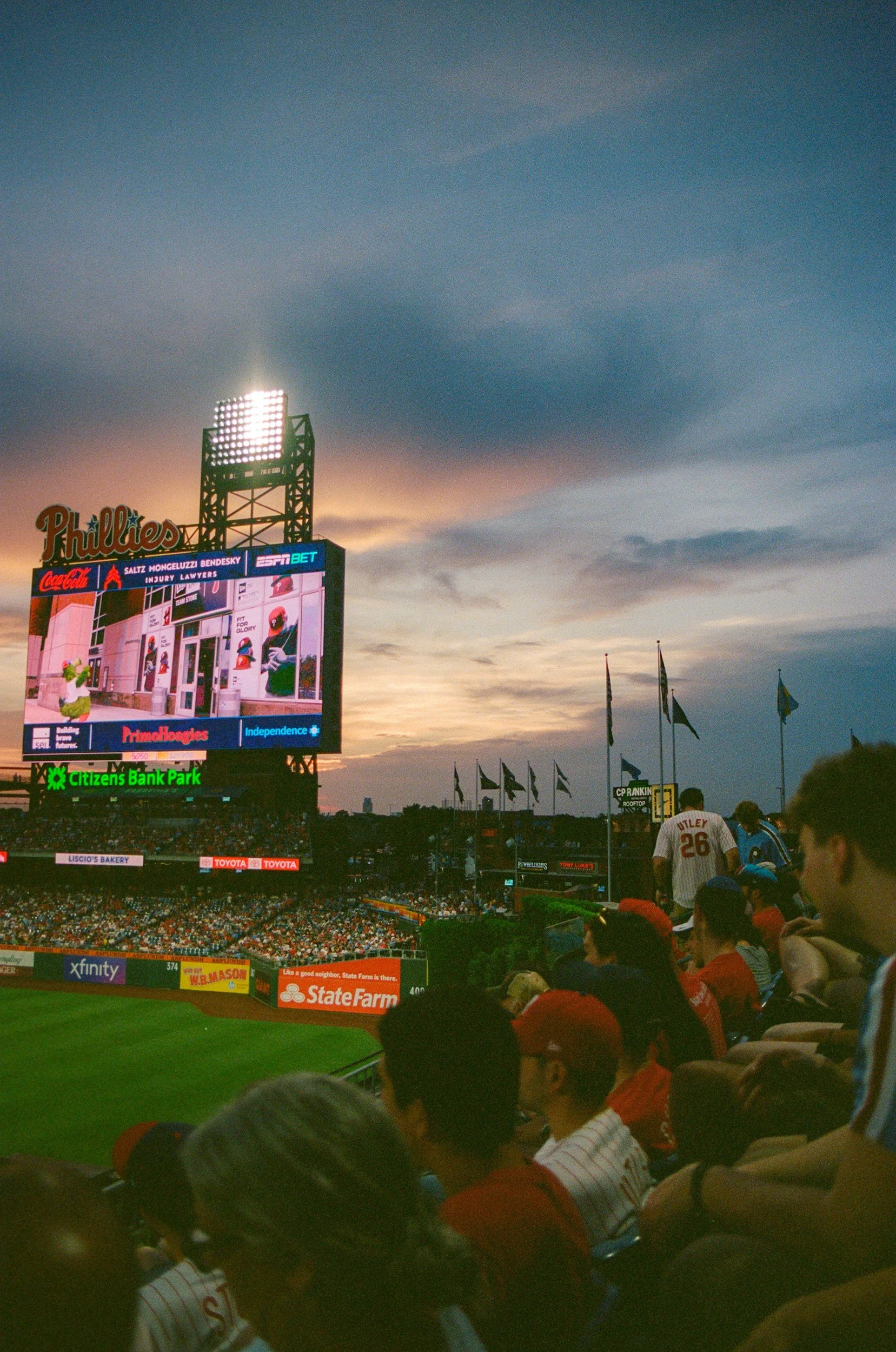 Crowd of baseball fans seated in Citizens Bank Park during a game at sunset, with a large video screen displaying advertisements and the Philadelphia Phillies logo.