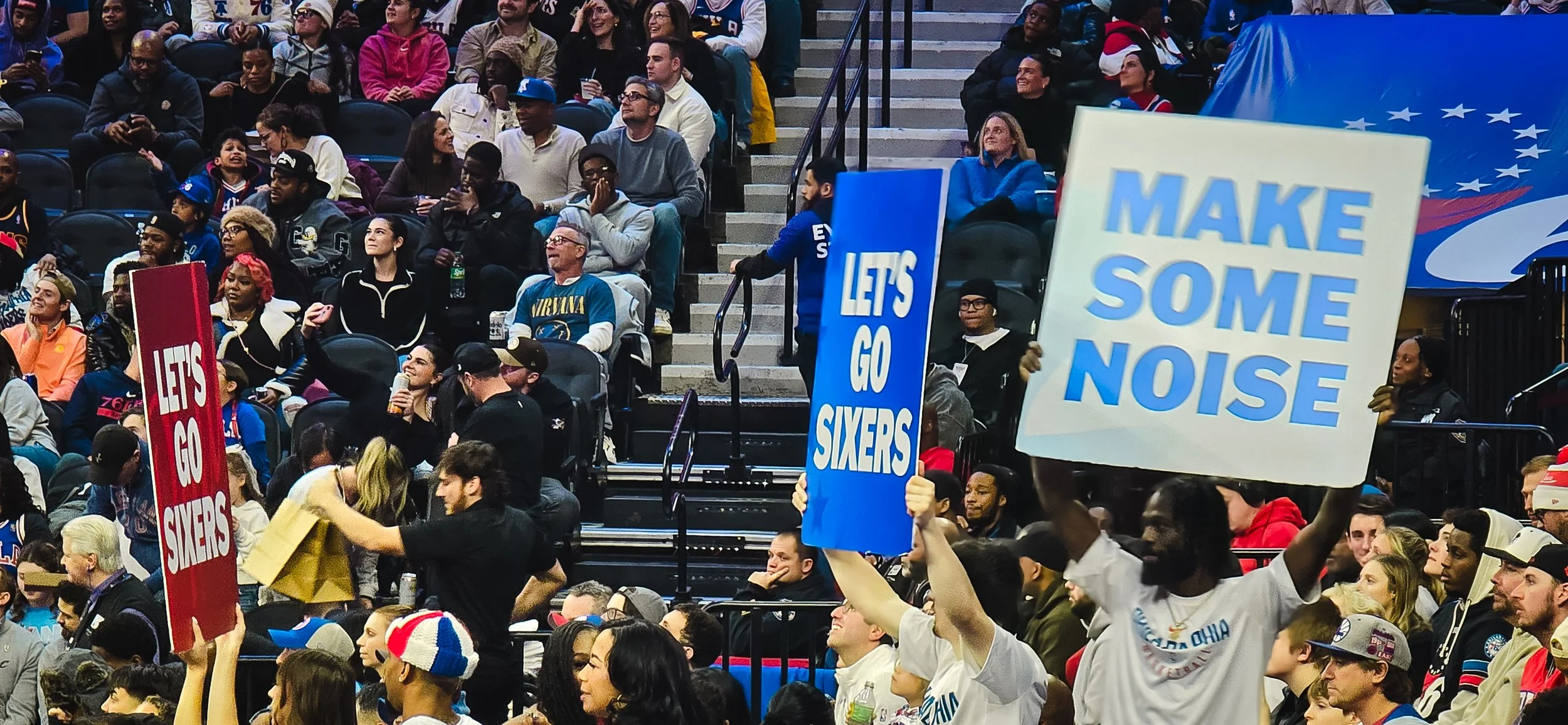 Crowd at a sports game with fans holding signs that say 'Let's go Sixers,' 'Make some noise,' and 'Let's go Sixers' in an indoor arena.