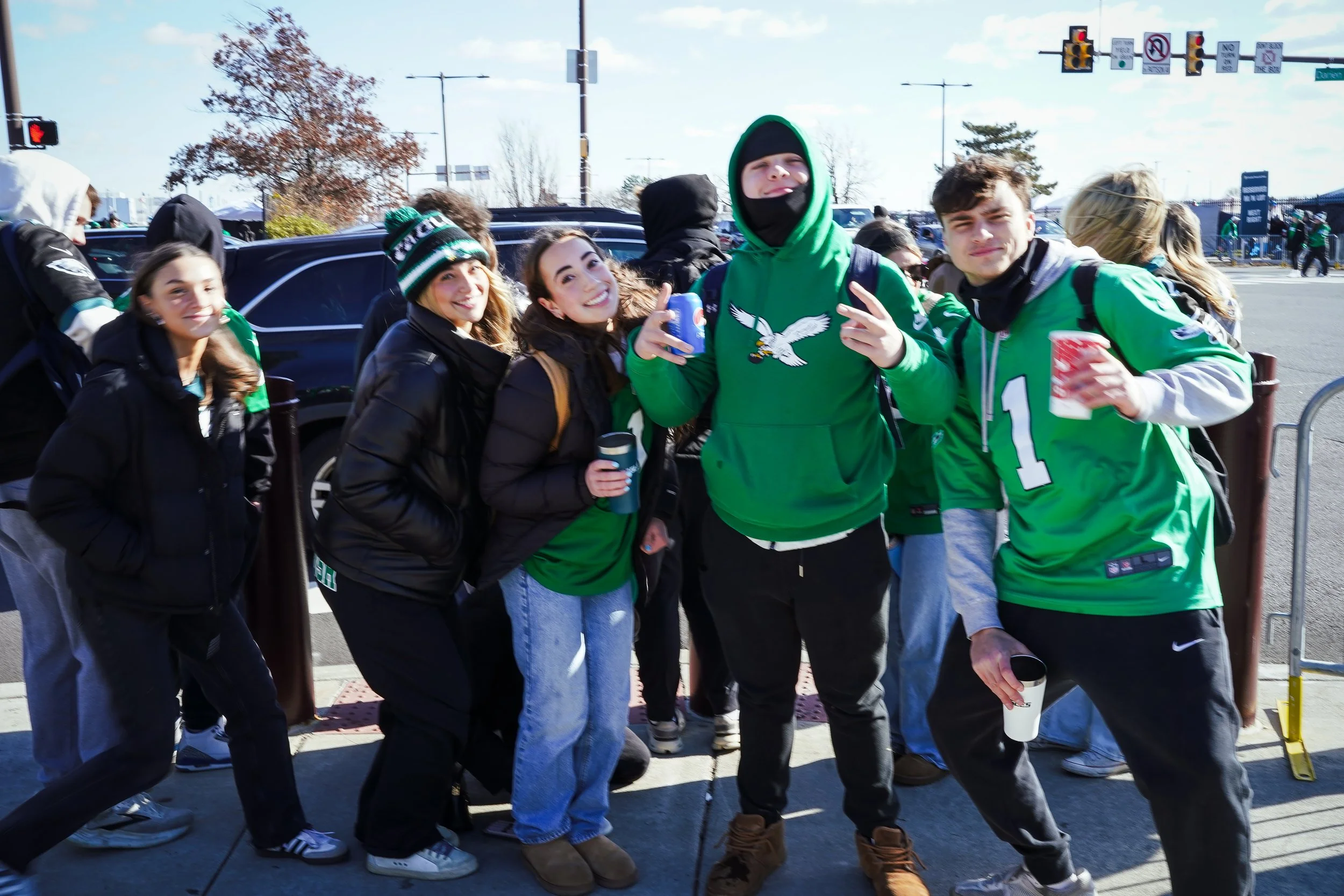 Group of young people wearing Philadelphia Eagles clothing, standing in a parking lot outdoors during daytime, some holding cups and making gestures.