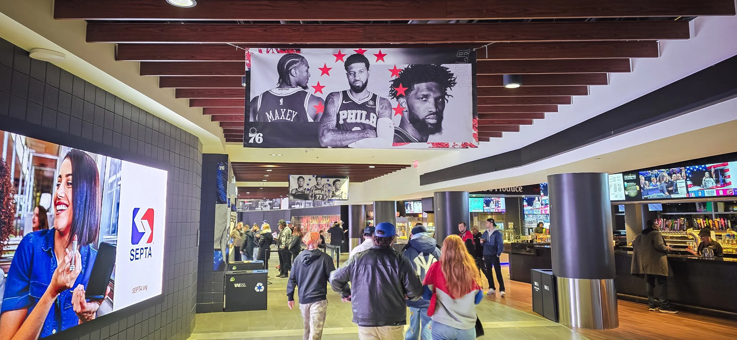 Indoor scene at a transit station with people walking and ordering food. Large digital screens display advertisements and news. Posters of basketball players hanging from the ceiling.