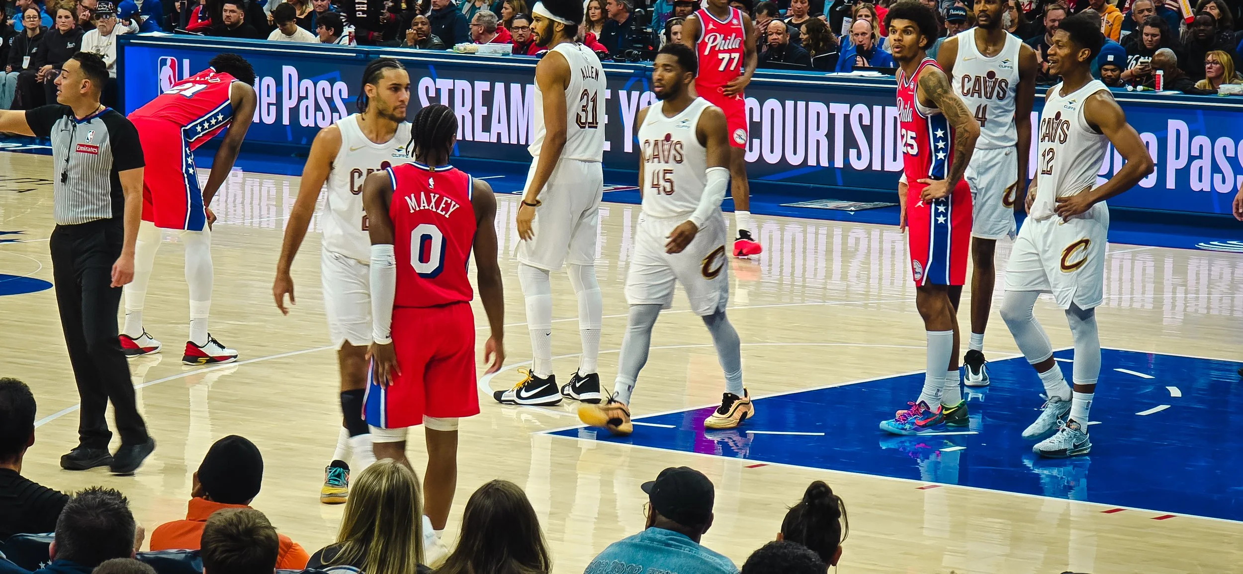 Basketball players on the court during a game, some wearing white jerseys of the Cleveland Cavaliers and others in red jerseys, with spectators watching from the stands.