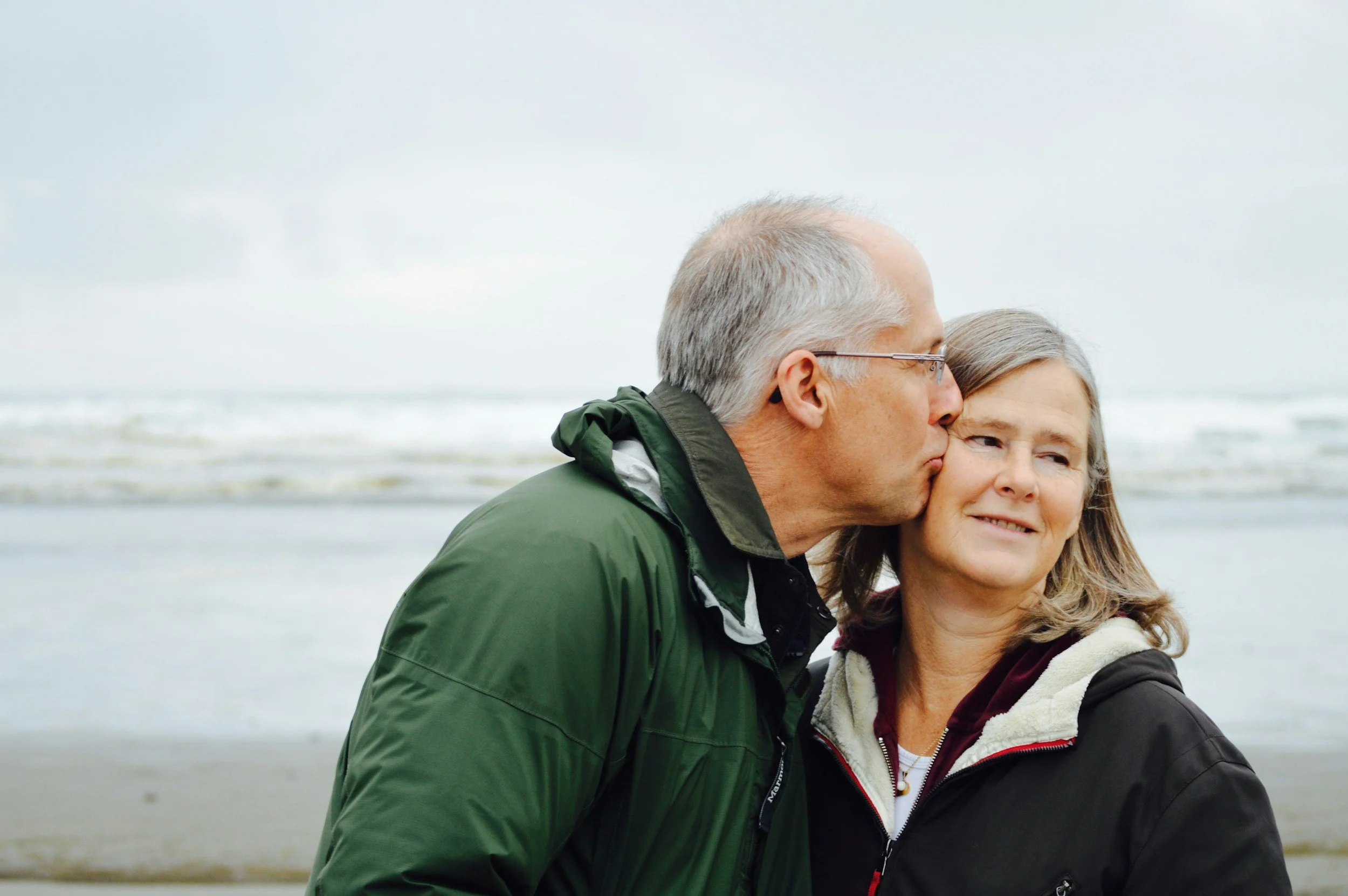 An elderly man with gray hair and glasses kisses an elderly woman with gray hair on the forehead as they stand on a beach with a cloudy sky and ocean waves in the background.