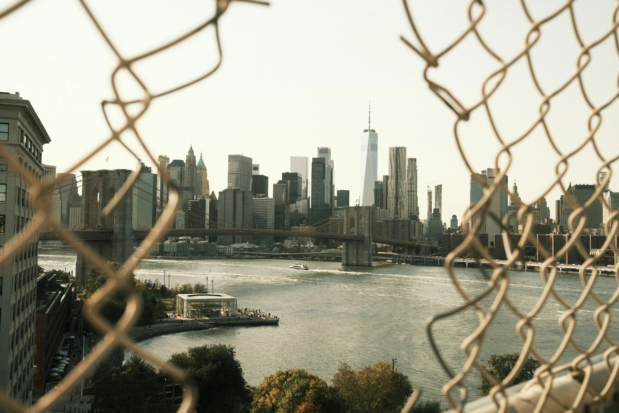 View of the Manhattan skyline through a chain-link fence, including the Brooklyn Bridge and the One World Trade Center.