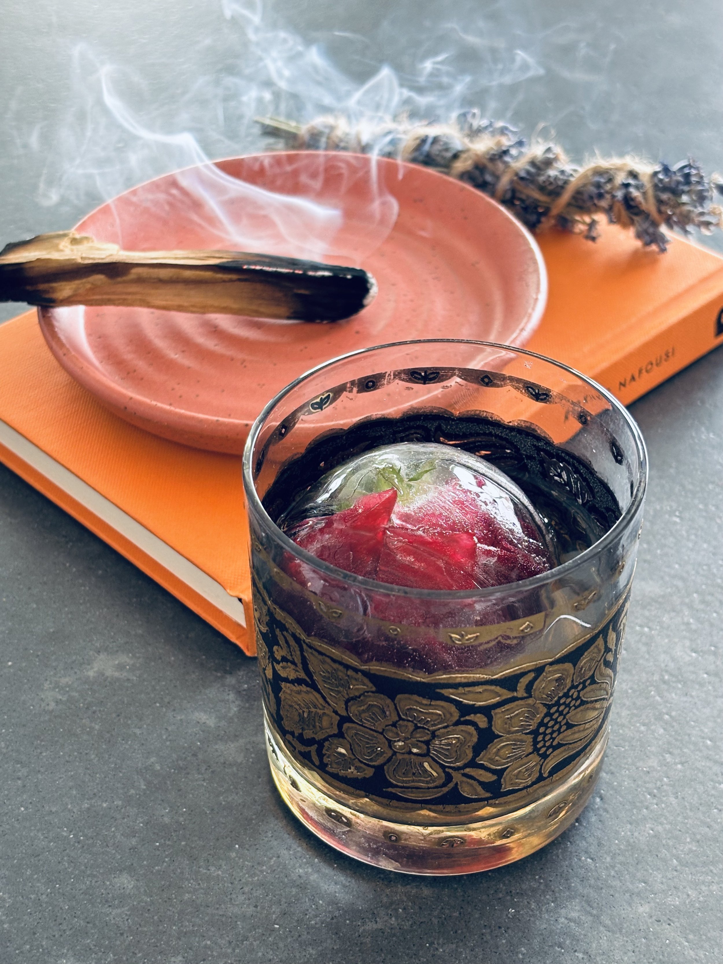 A social media drink in a decorative glass with a floral pattern, contains ice and a red liquid. Behind it, a pink plate with a burnt sage stick resting on it, emitting smoke. There is also a bundle of dried sage or similar herb on an orange book or box.