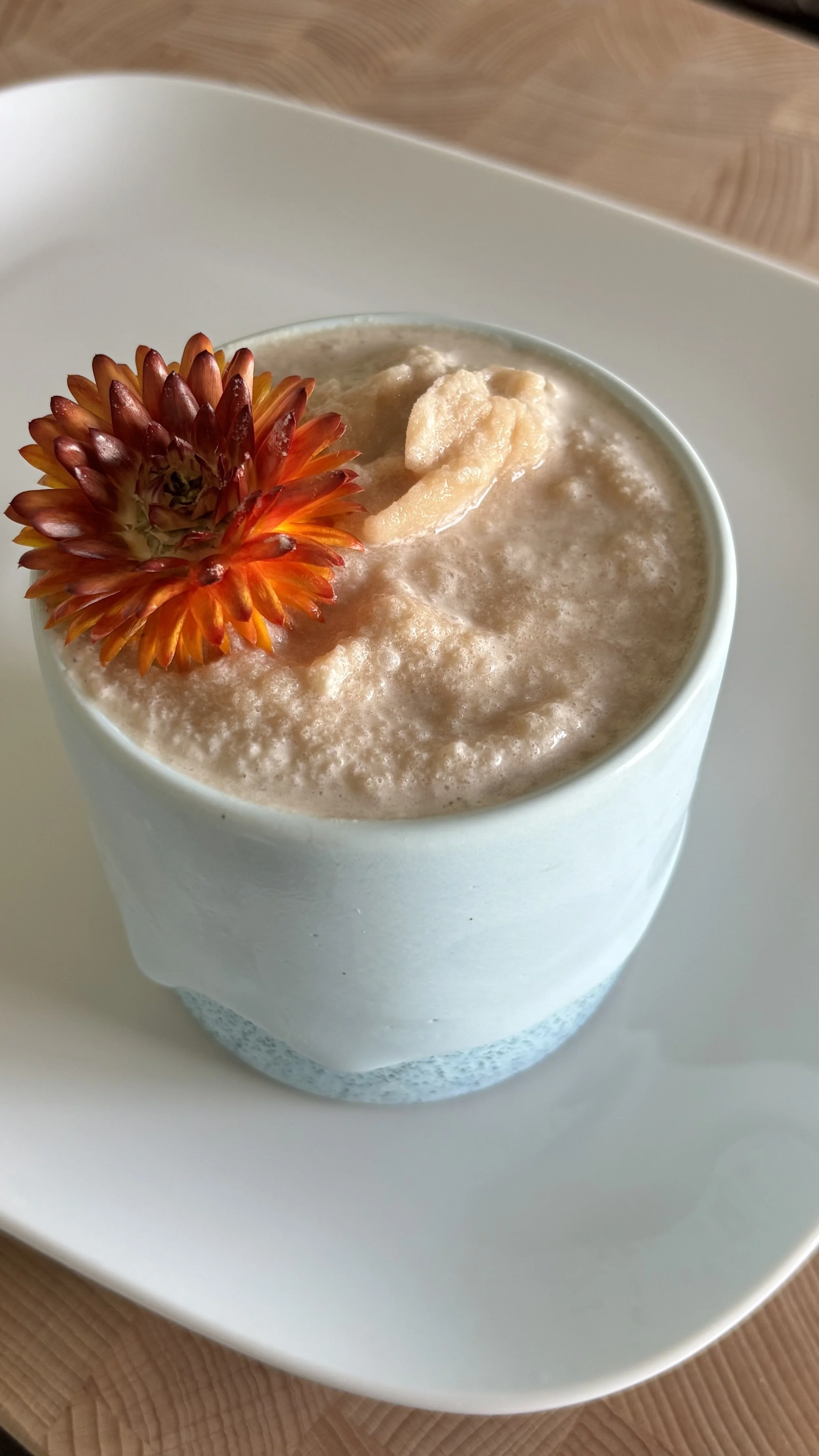 A white ceramic cup filled with a frothy, beige-colored beverage, garnished with an orange and red flower and a small piece of fruit or other topping, on a white square plate resting on a wooden surface.