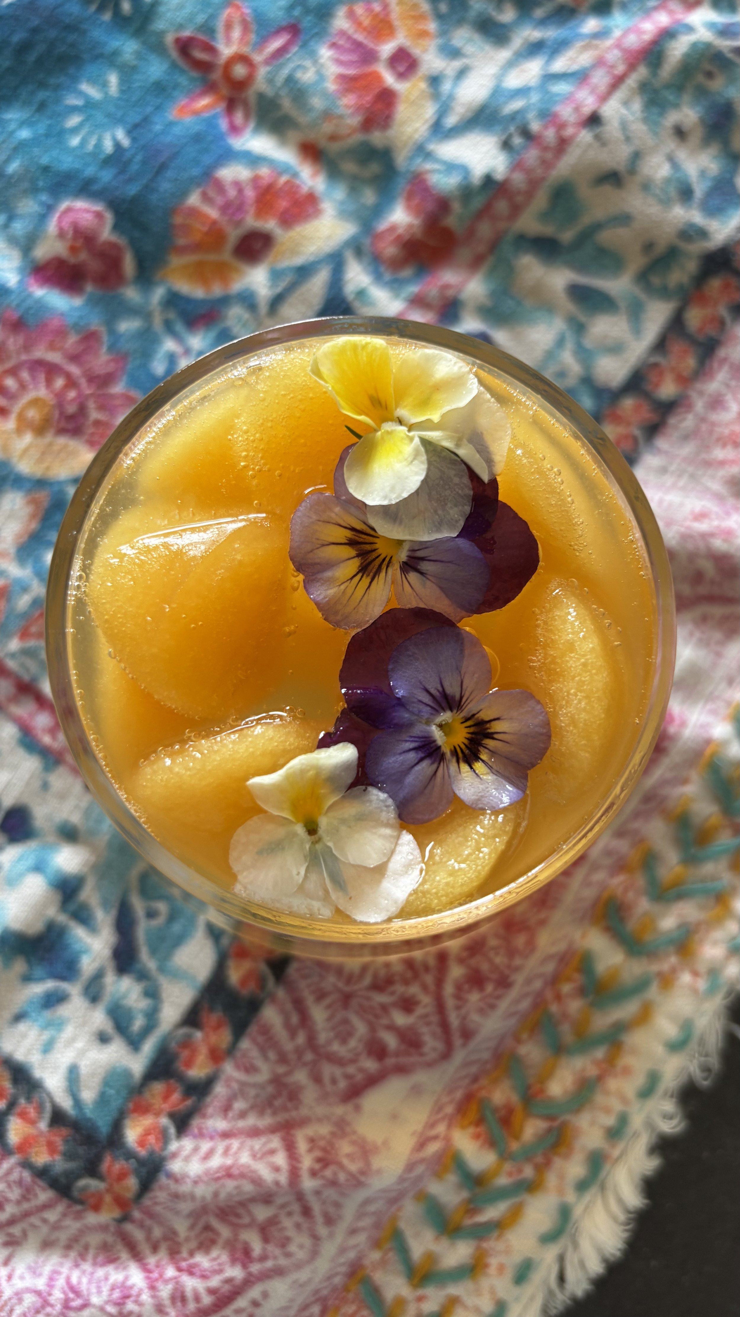 A glass of yellow-orange drink garnished with colorful edible flowers, placed on a multicolored fabric with floral and geometric patterns.