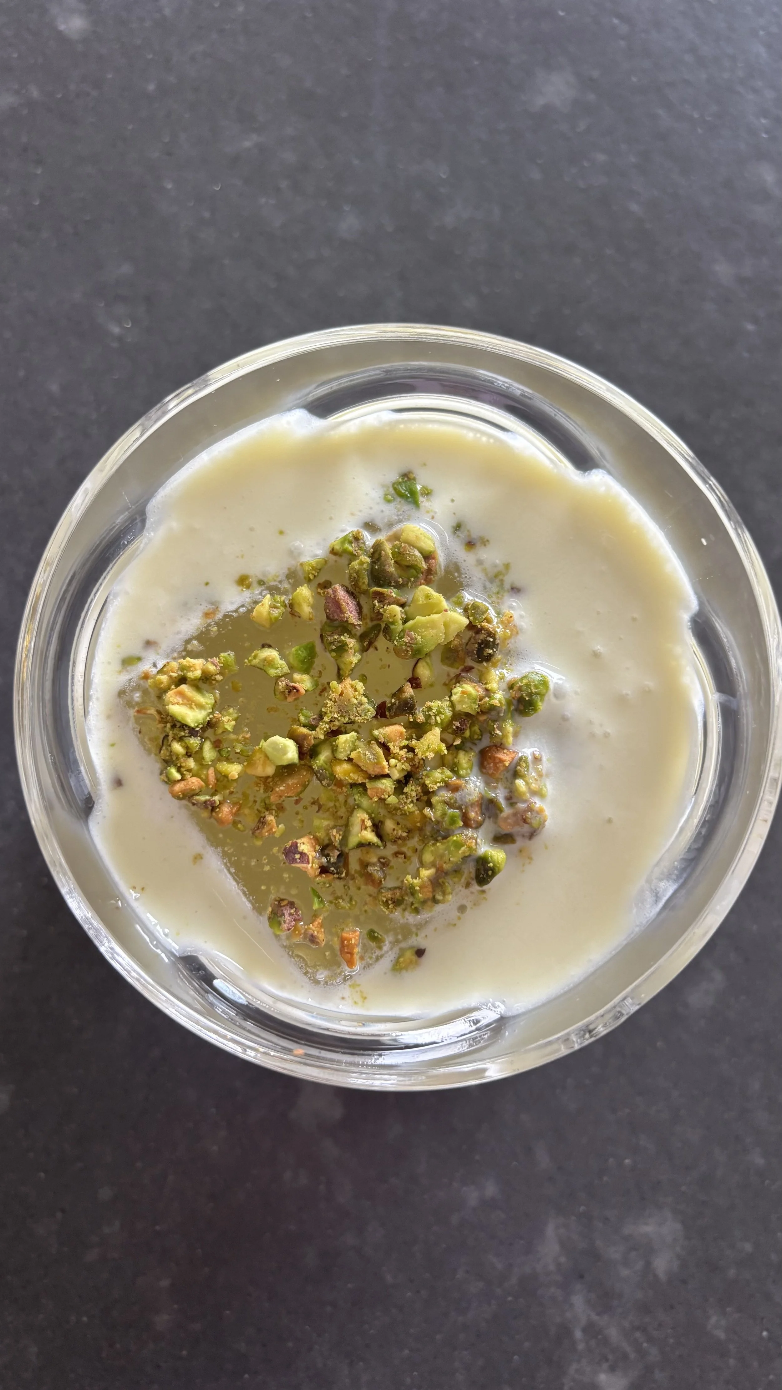 A clear glass bowl of pistachio pudding topped with chopped pistach nuts on a black surface.