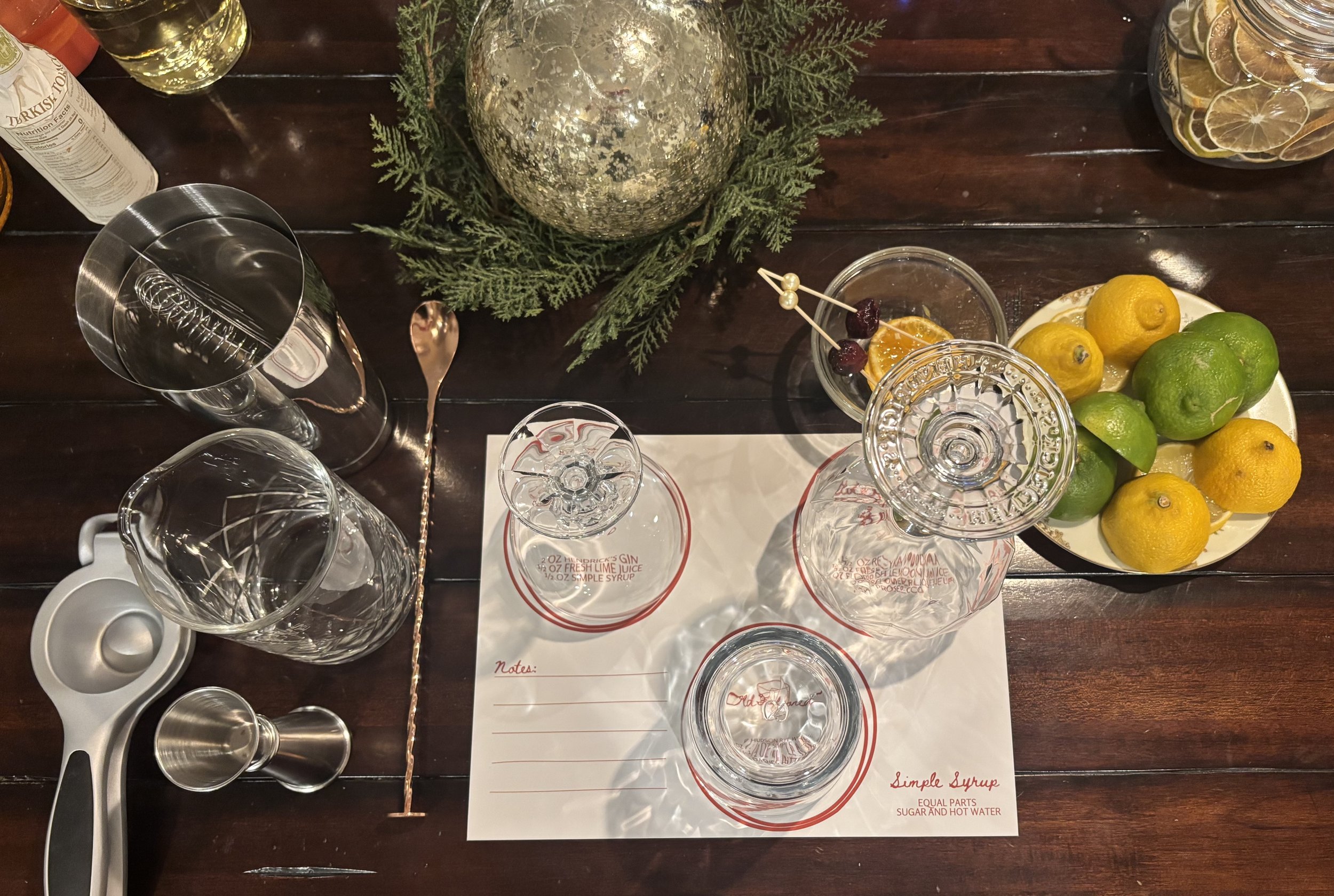 A top-down view of a bar or cocktail mixing setup on a wooden table, including empty glasses, a tray with lemons and limes, a glass of citrus garnished with cherries, a metal shaker, a muddler, and a notepad with simple syrup instructions, with some greenery and a decorative globe in the background.