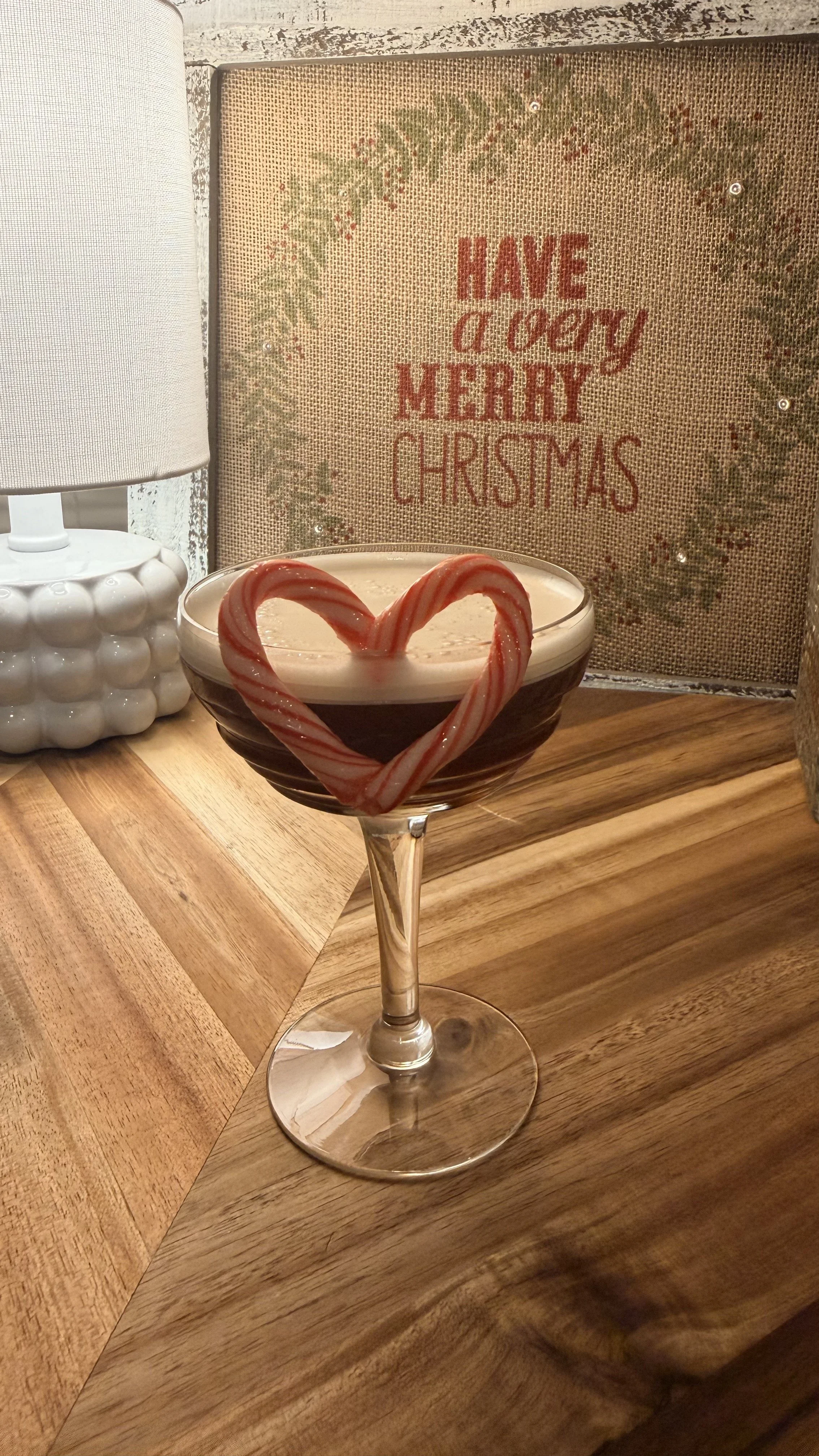 A festive Christmas drink in a glass with a candy cane heart decoration on the rim. In the background, a rustic sign with the message "Have a very Merry Christmas" and a white lamp with a textured base.