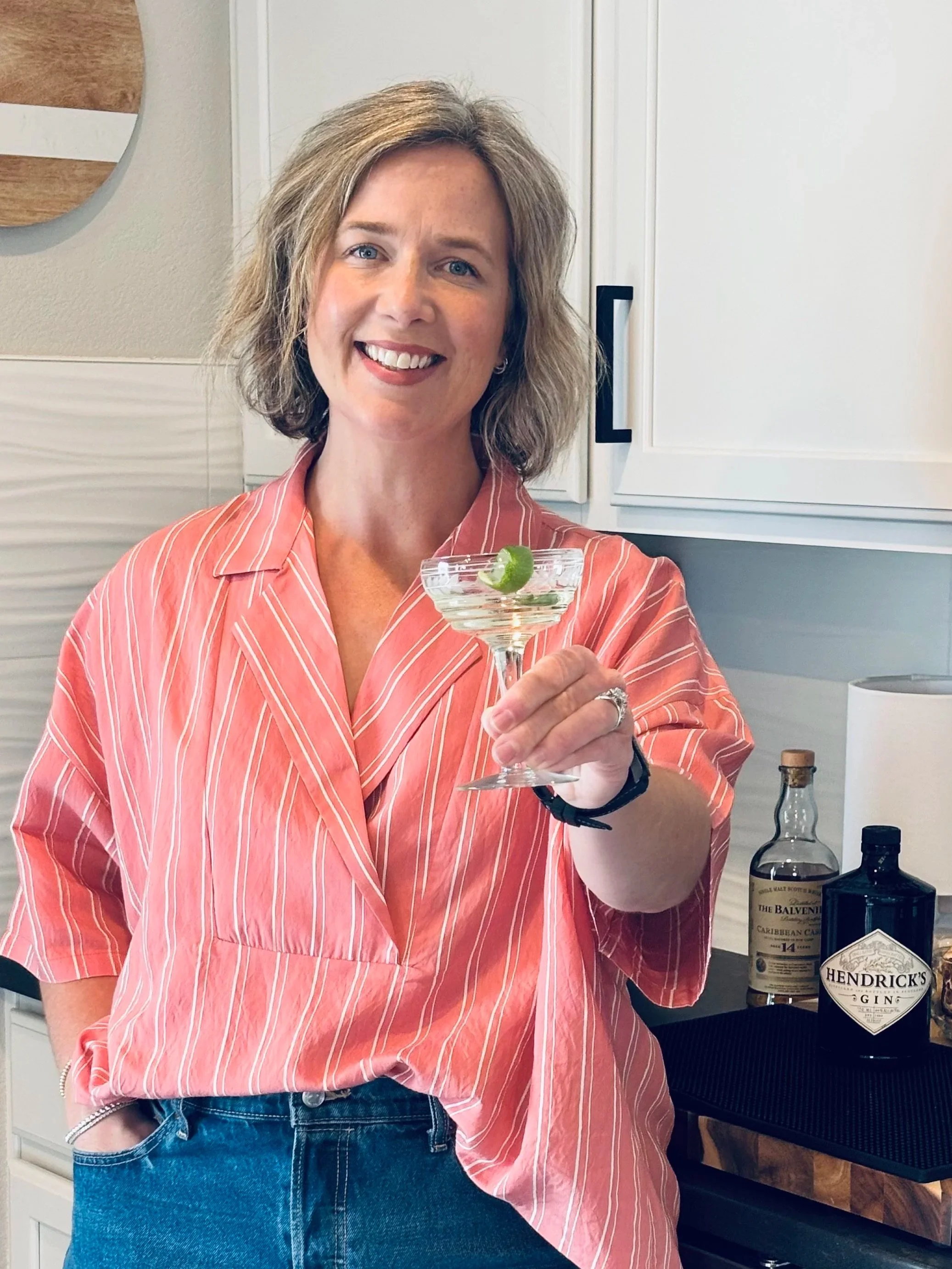 A woman with short blonde hair smiling and holding a cocktail with a lime garnish, standing in a modern kitchen.