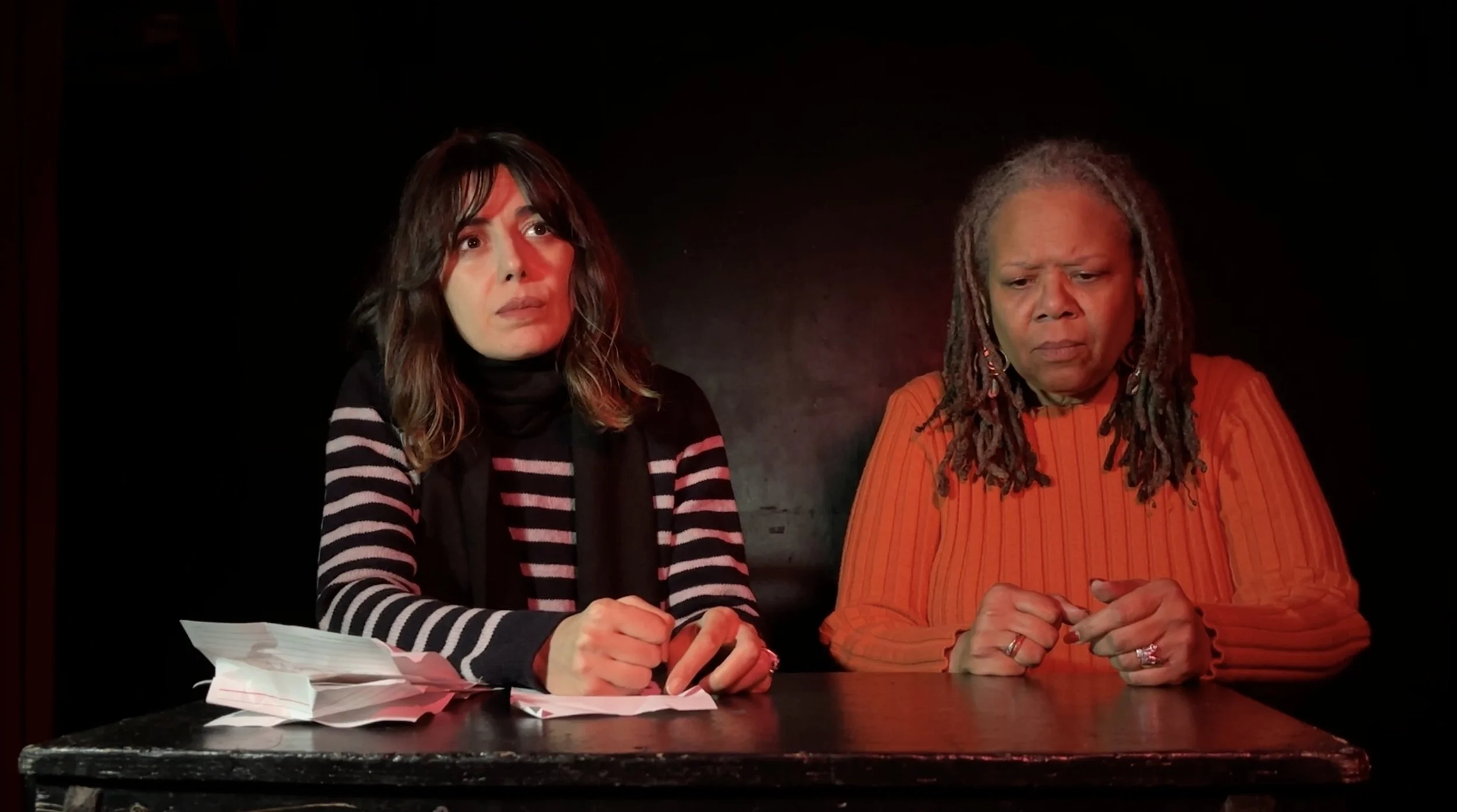 Two women seated at a table in a dark room; woman on left has shoulder-length brown hair, wearing a black and white striped sweater; woman on right has long gray dreadlocks, wearing an orange striped shirt; both look serious or concerned, with papers on the table.