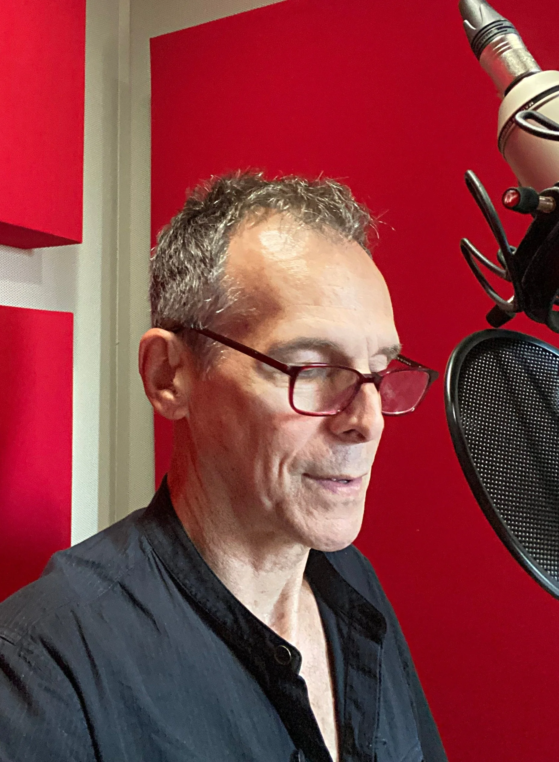 A man with red glasses is speaking into a professional microphone in a studio with red and gray walls. This is T. Ryder Smith director and designer of Liberty Scrap by Christina Masciotti premiering at Culture Lab LIC in March 2026.