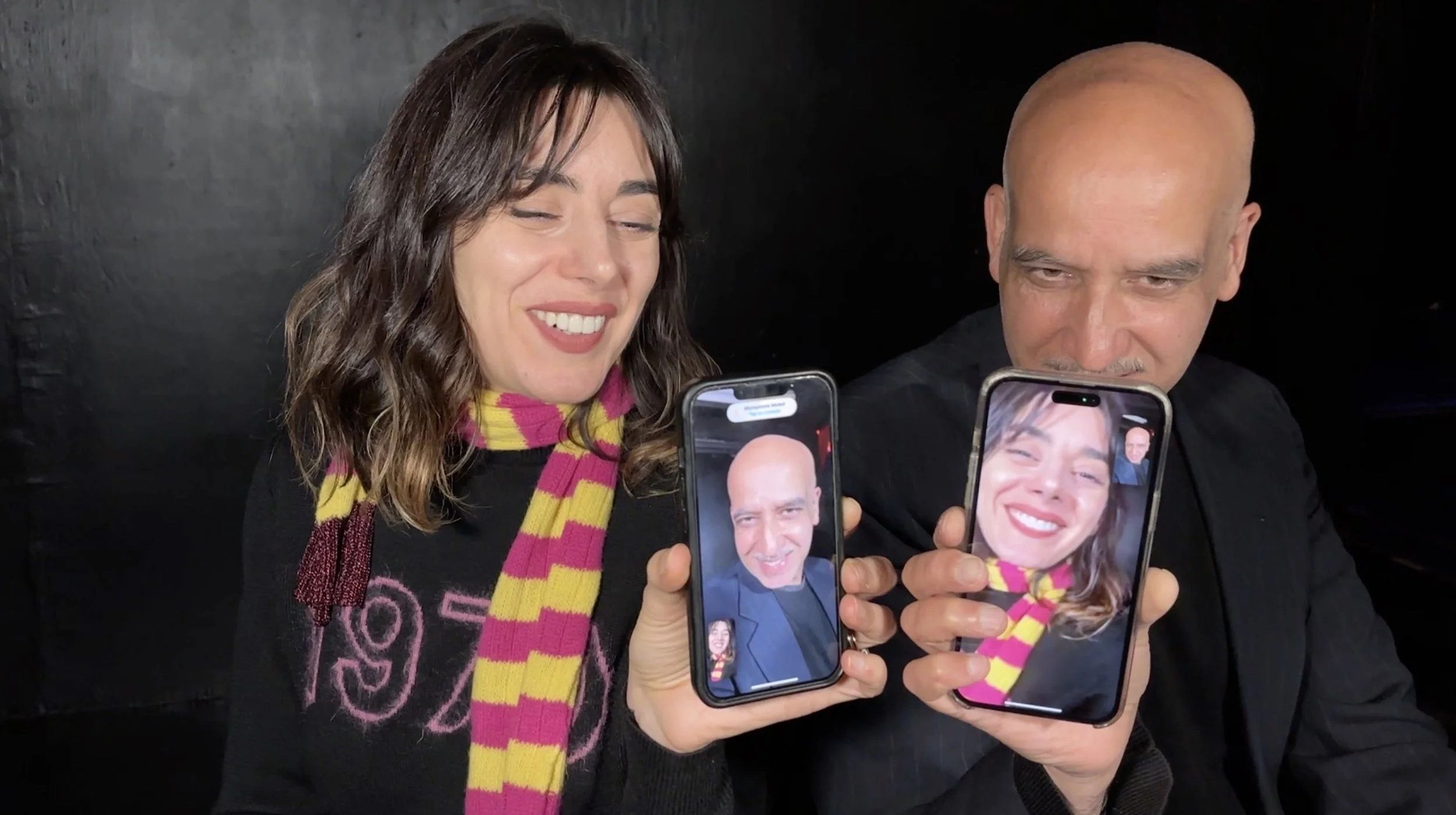 Two people are smiling and showing their smartphones, Natia Dune as Katya, and Alok Tewari as Hidayat in LIBERTY SCRAP a new play by Christina Masciotti, directed and designed by T. Ryder Smith at Culture Lab LIC in March 2026.