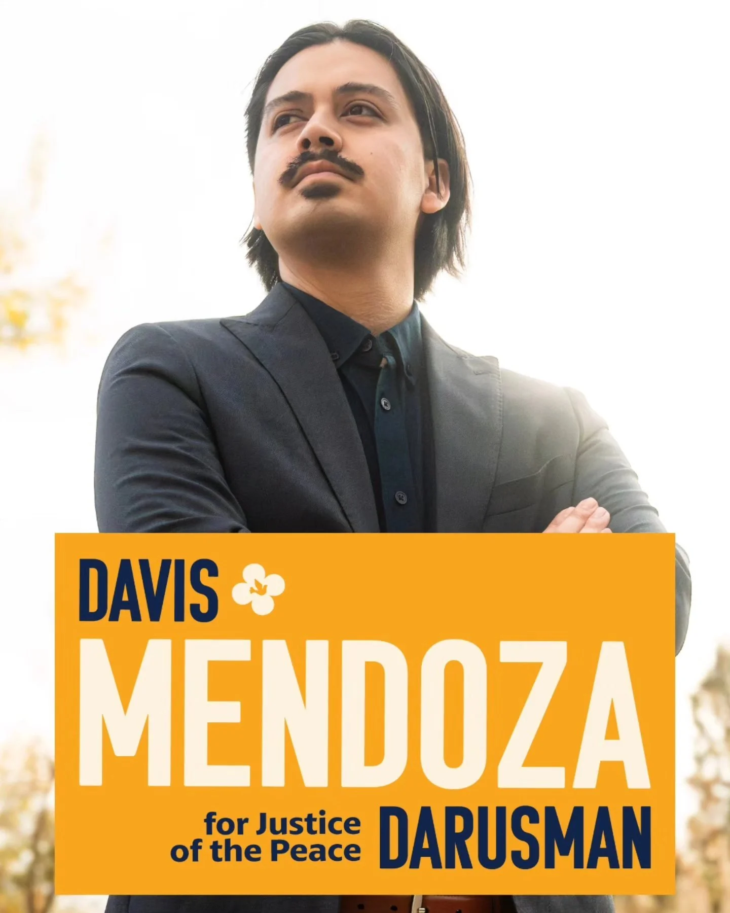 A man with dark hair and a mustache wearing a dark suit standing outdoors with arms crossed, with campaign text overlayed that reads: 'Davis Mendoza for Justice of the Peace Darusman' with a small flower icon.