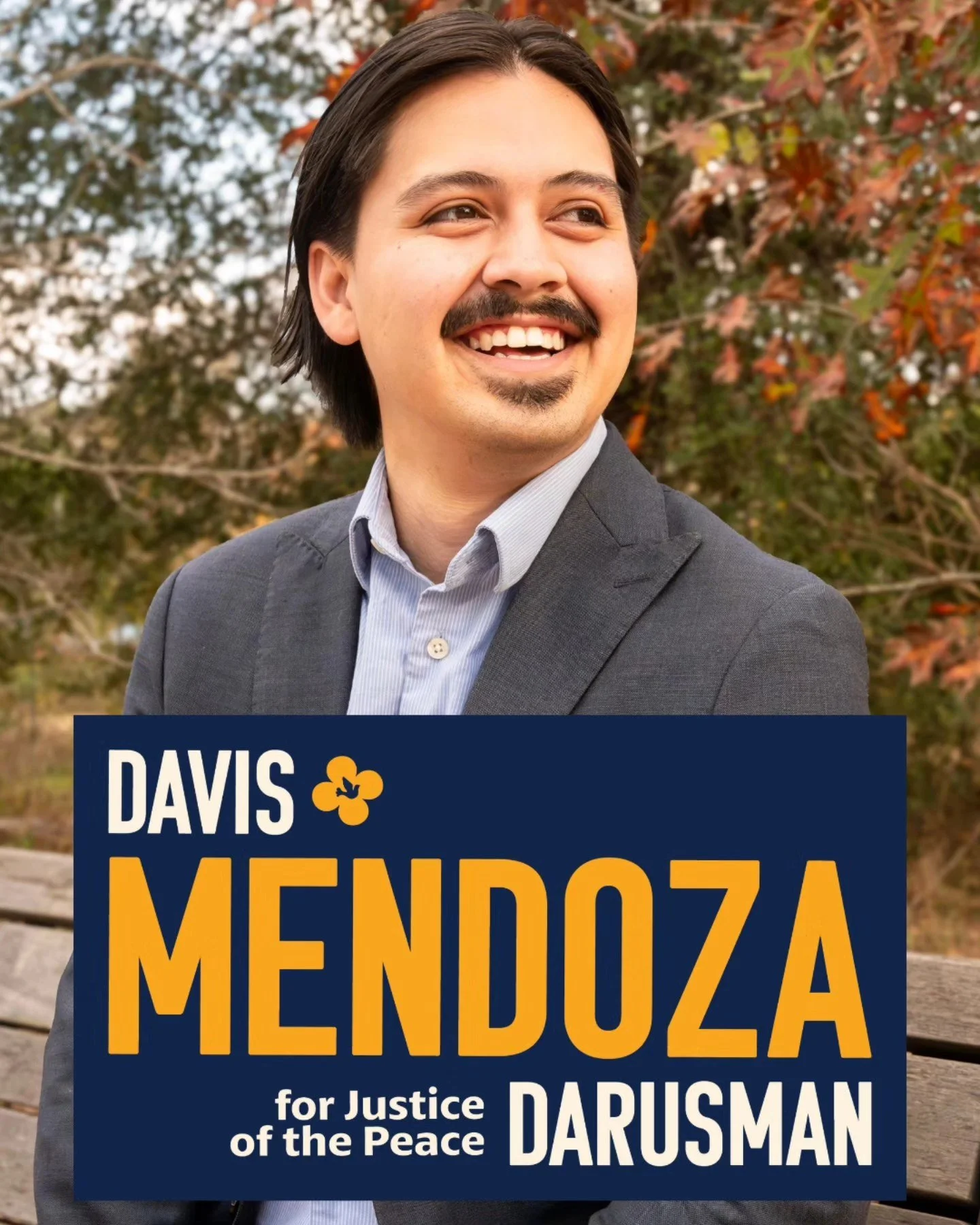 A man with dark hair and a mustache, smiling and looking to the side, wearing a gray suit jacket and shirt, sitting on a park bench with autumn foliage in the background. A political campaign sign in front reads "Davis Mendoza for Justice of the Peace Darusman."