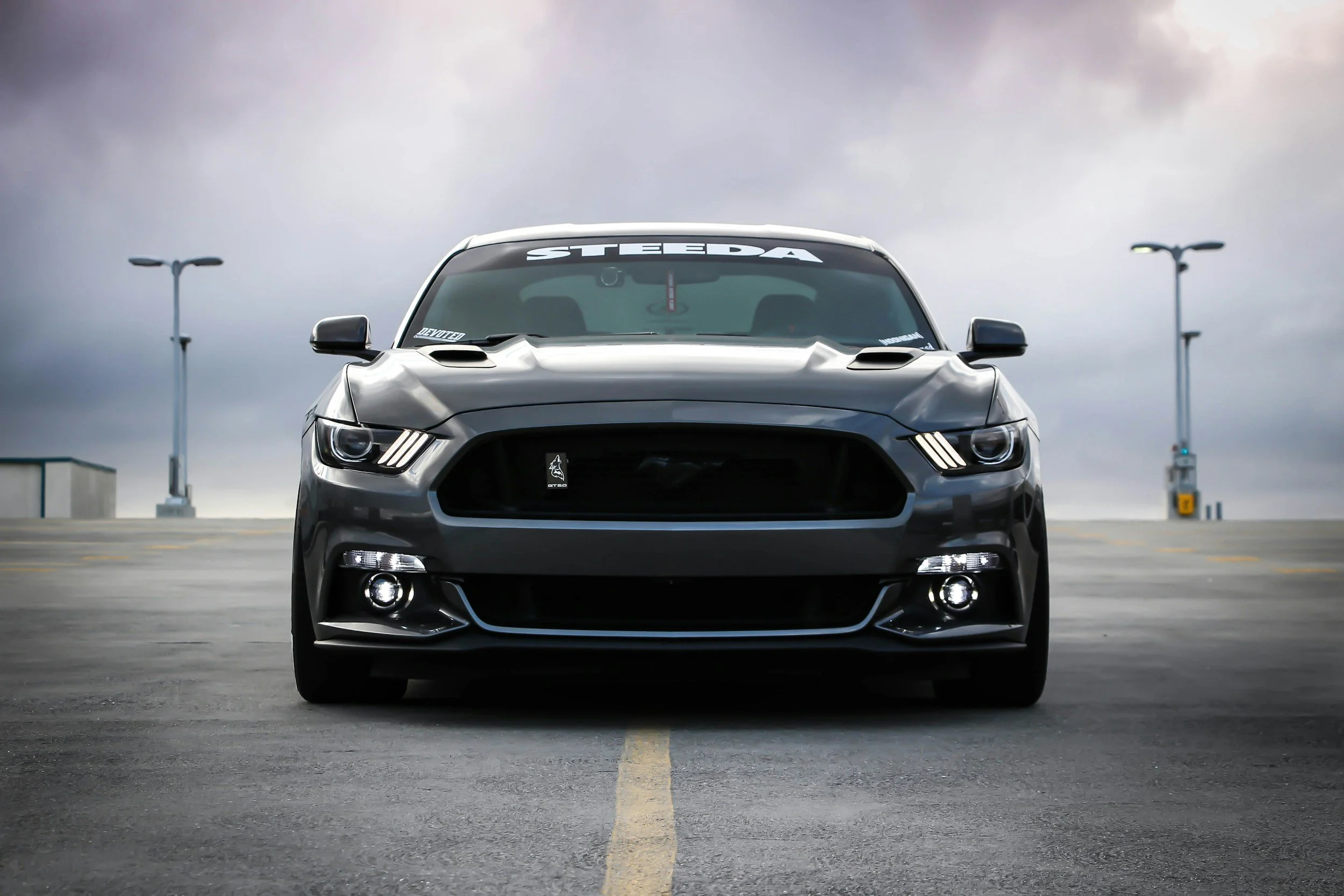 Front view of a gray Ford Mustang parked in an empty parking lot during overcast weather, with streetlights in the background.