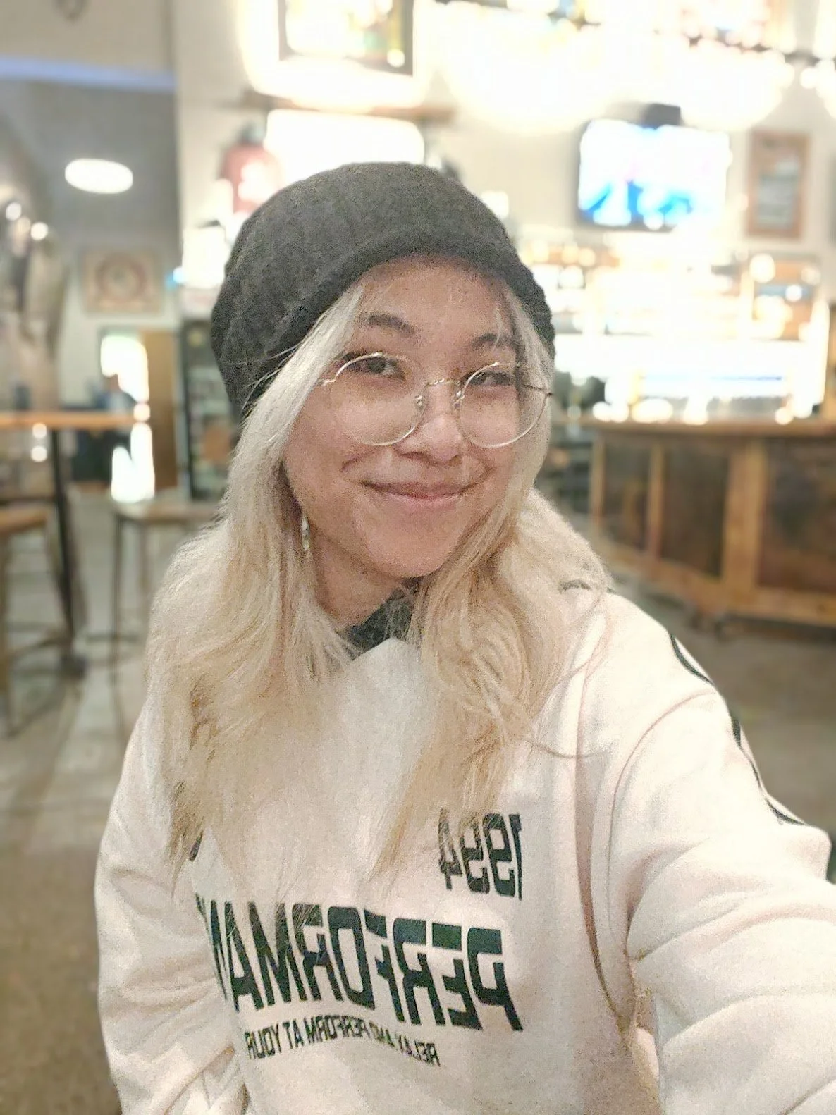 A woman with blonde hair wearing glasses, a black beanie, and a white sweatshirt, smiling in a restaurant or cafe interior.