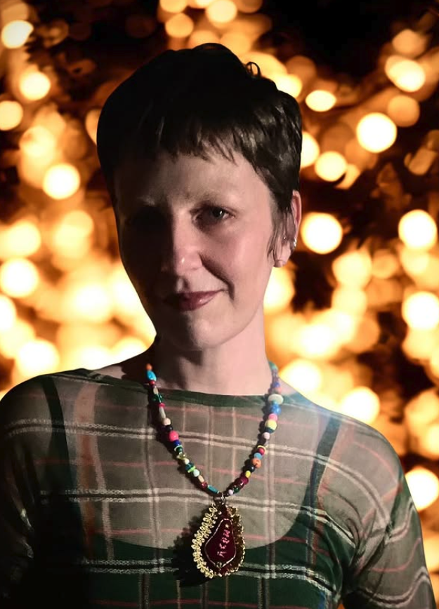 A woman with short dark hair wearing a plaid dress and a colorful beaded necklace with a large red pendant, standing in front of a background with warm, blurred lights.